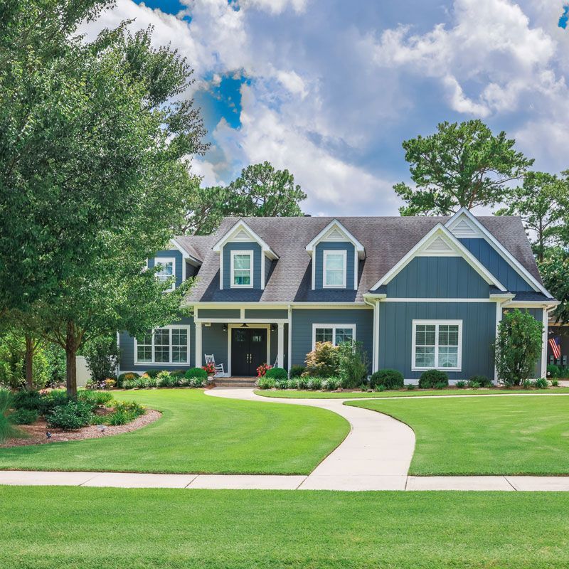 Blue suburban house with a manicured lawn, curved walkway, and trees under a partly cloudy sky