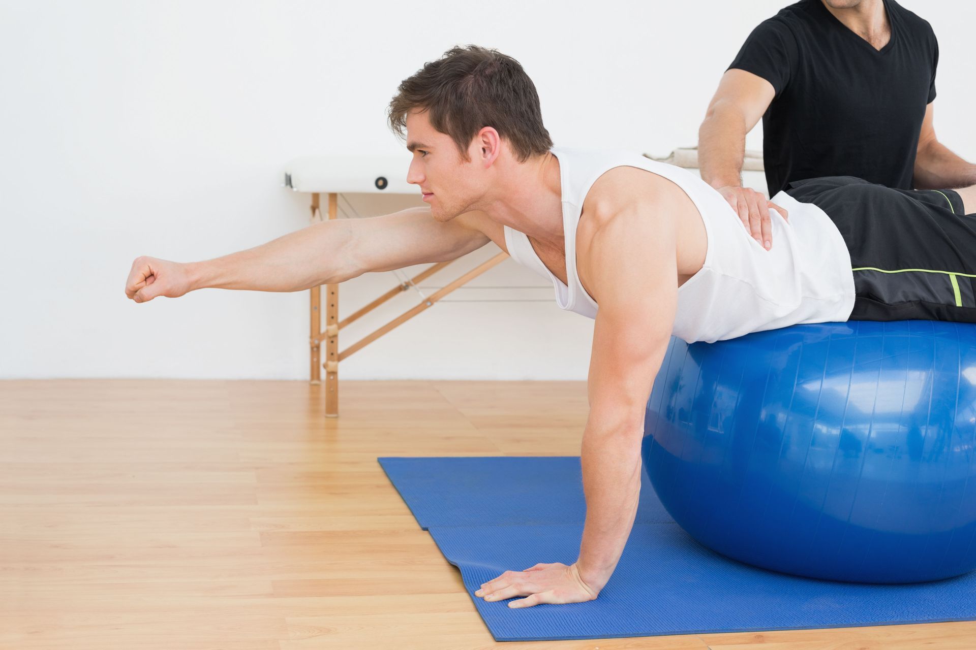 Man exercising on blue exercise ball, assisted by a person. Both in a gym setting.
