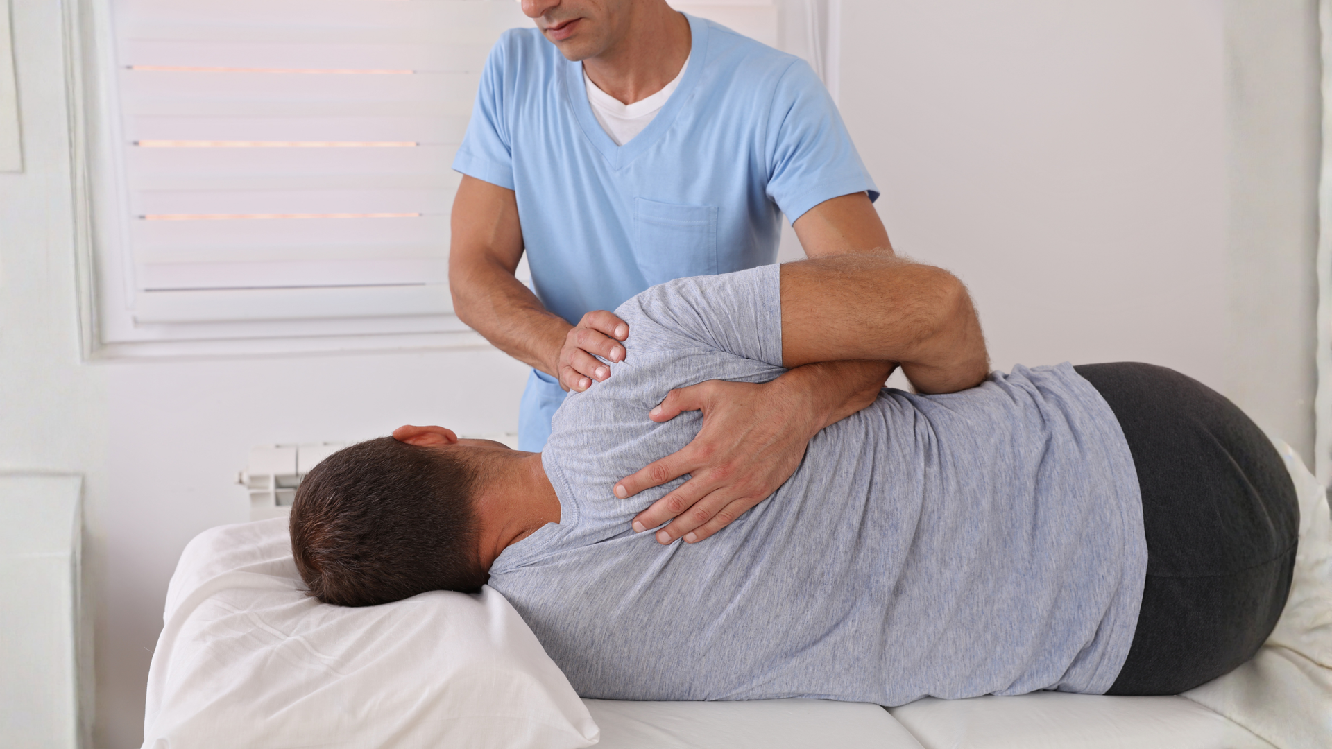 Man receiving physical therapy, lying on table while therapist stretches his shoulder.