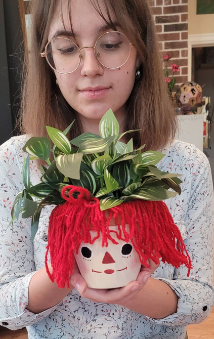 A woman is holding a potted plant with a doll face on it.