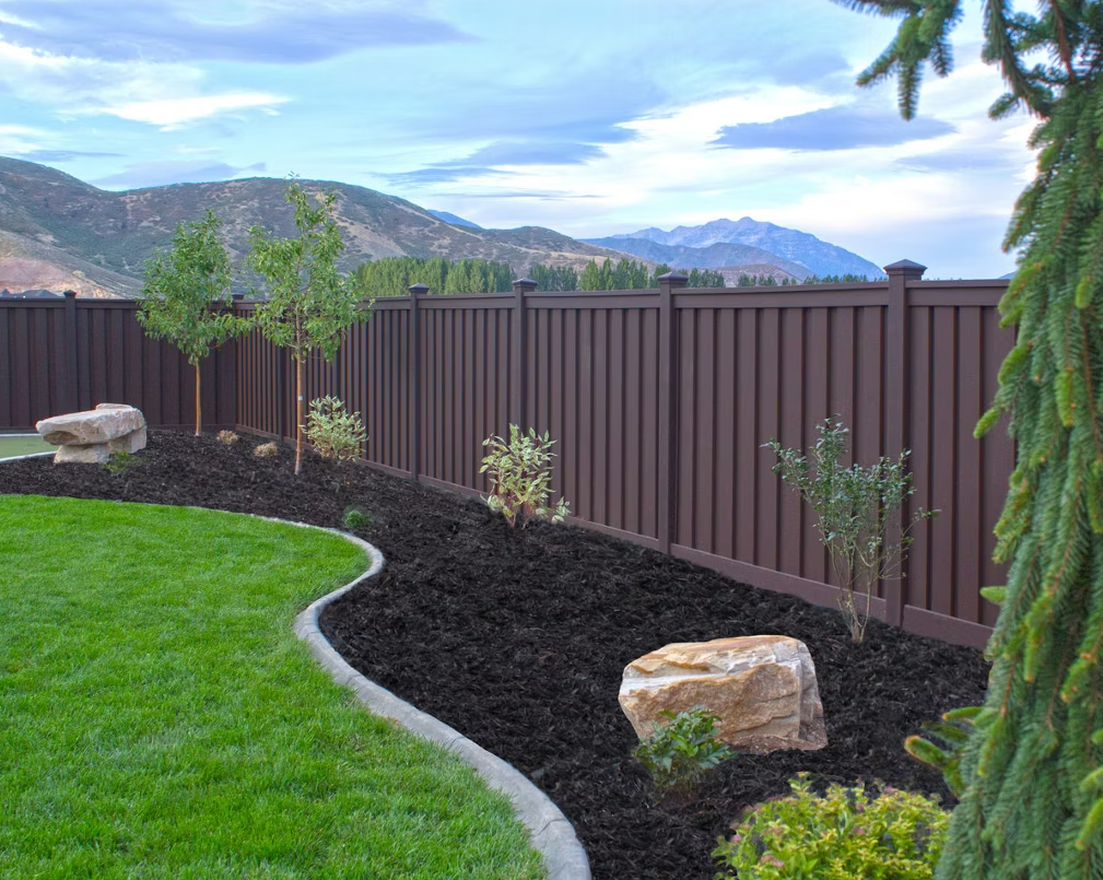Brown vertical-slat fence bordering a lawn with dark mulch, small trees, and a mountain backdrop.