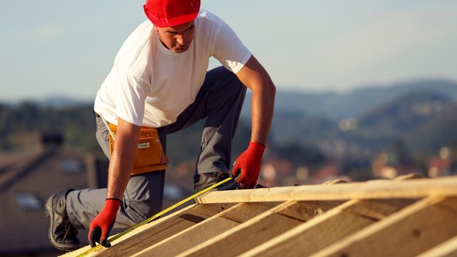 Carpenter on a rooftop, measuring wood framing with a tape measure. Wearing a red hard hat and gloves.