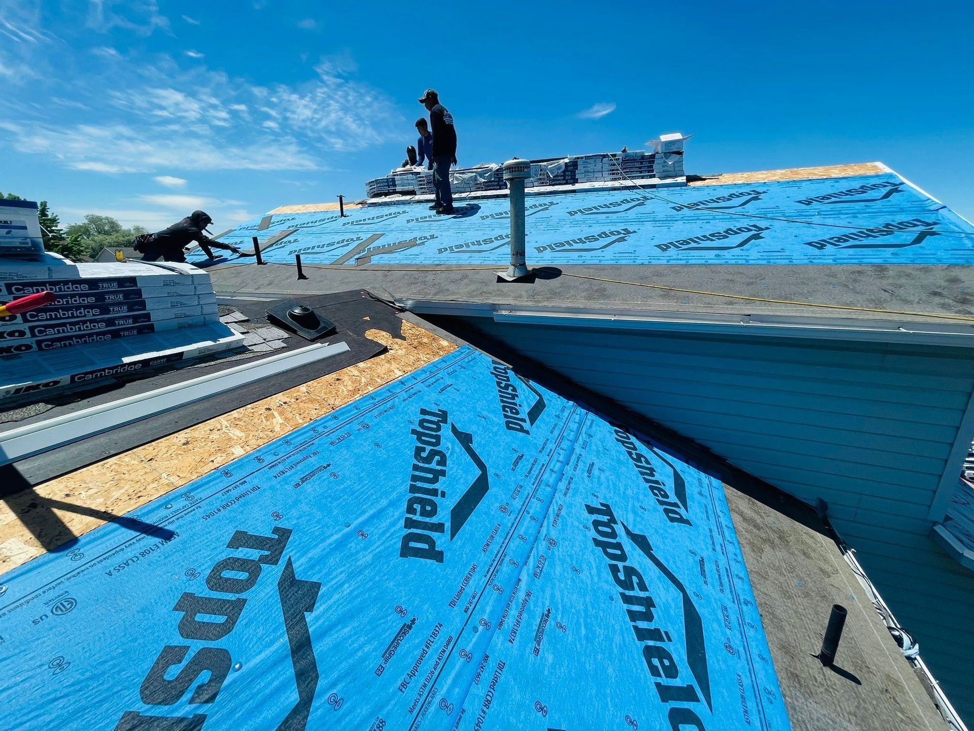 Roofers installing shingles on a house roof under a sunny blue sky, using blue underlayment.