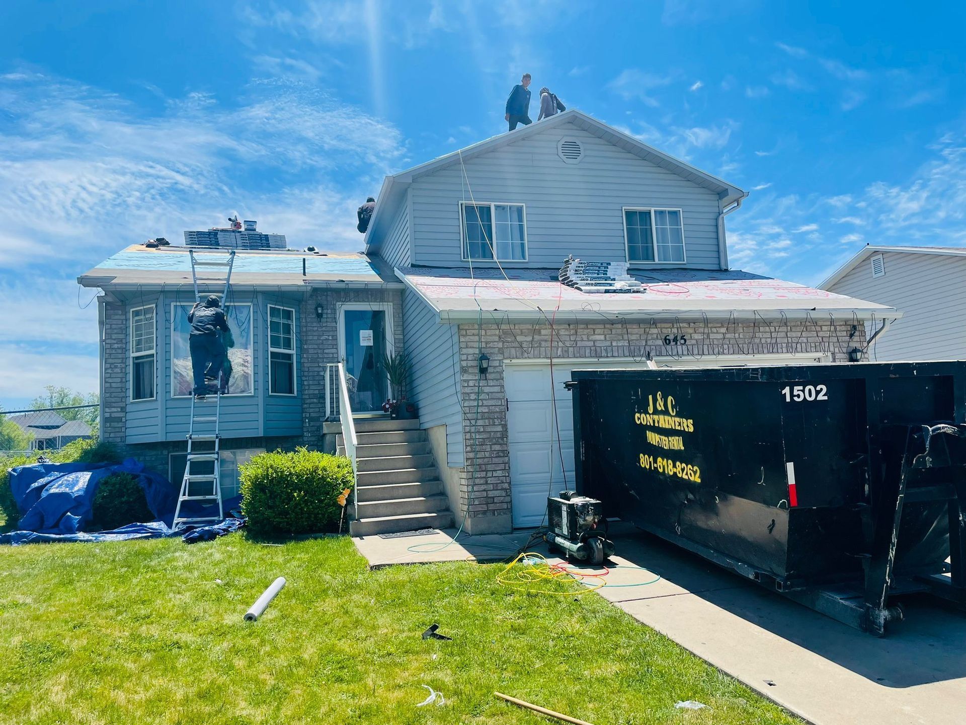 Riverbend workers on a house roof replacing shingles on a sunny day in Brigham City. A dumpster sits in the driveway.