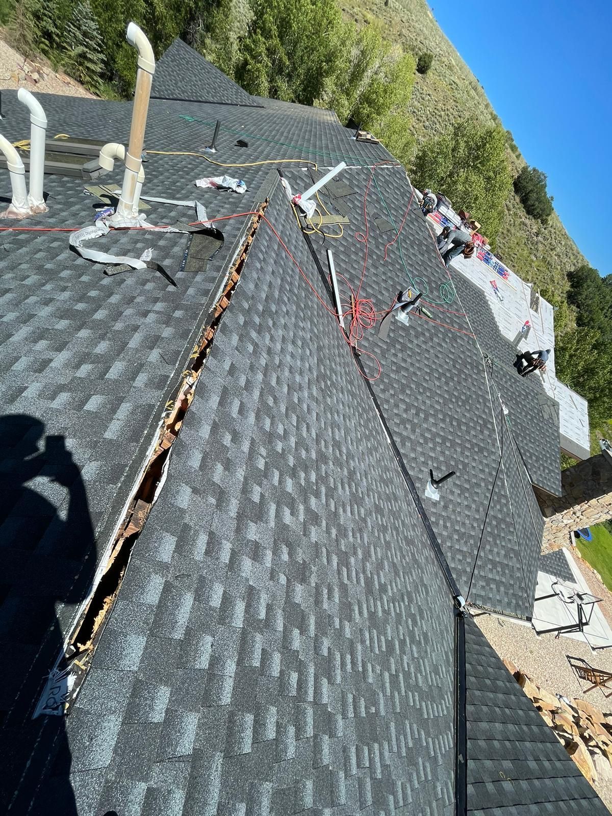 View of a shingled roof with damaged wood, chimneys, and wires; a hillside backdrop in Idaho Falls, ID.