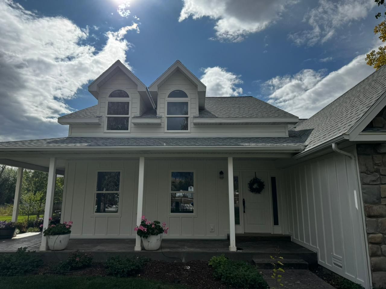White house with porch, dormers, and light gray roof under a cloudy blue sky.