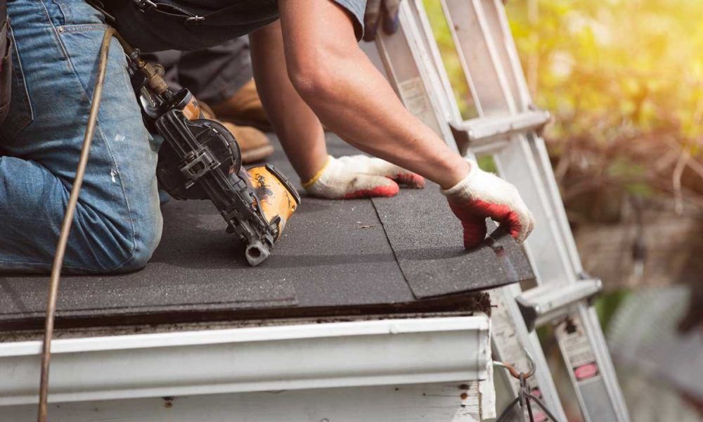 Person installing asphalt shingles on a roof, using a nail gun; ladder and gutter visible.
