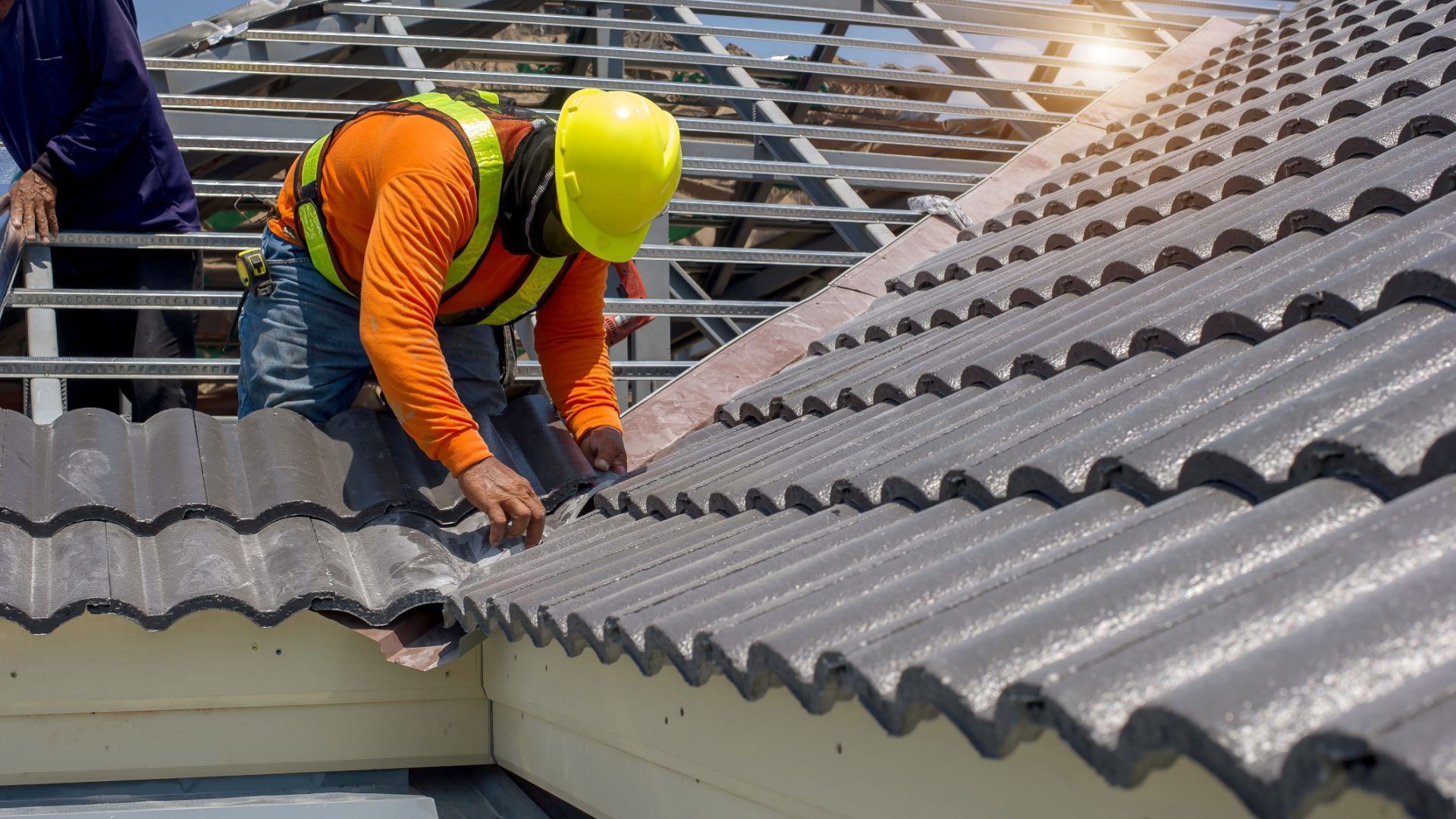 Roofer in orange shirt and yellow hard hat installing gray ceramic roof tiles on a house.