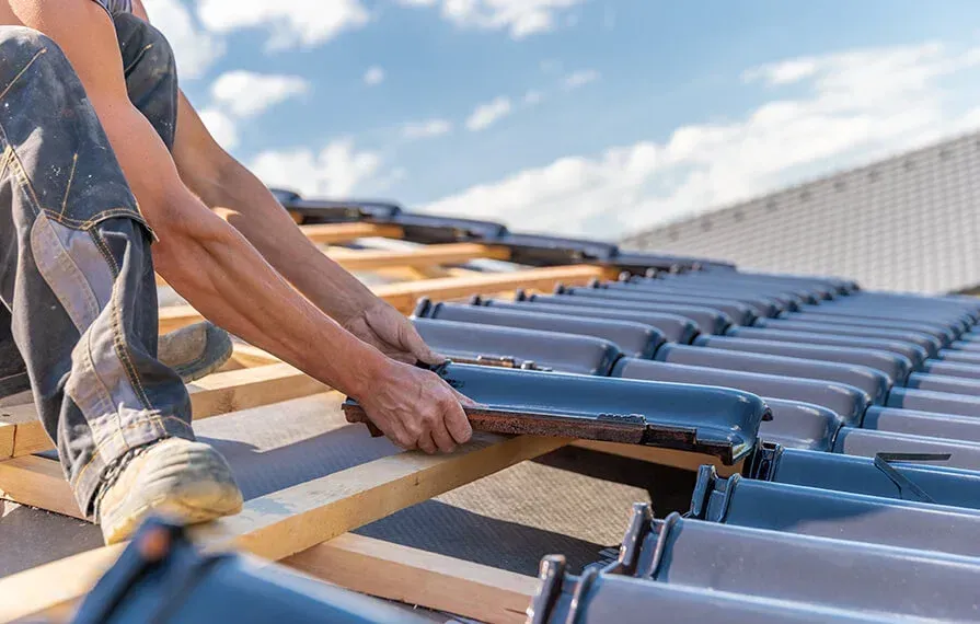 Roofer installing dark gray tiles on a wooden roof framework under a sunny sky.