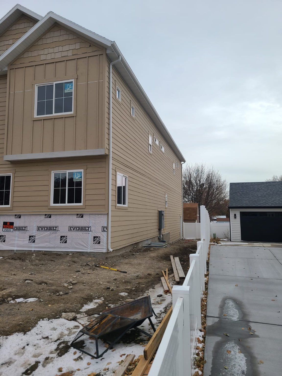 Two-story house with tan siding under construction, next to a white fence and a garage on a cloudy day.