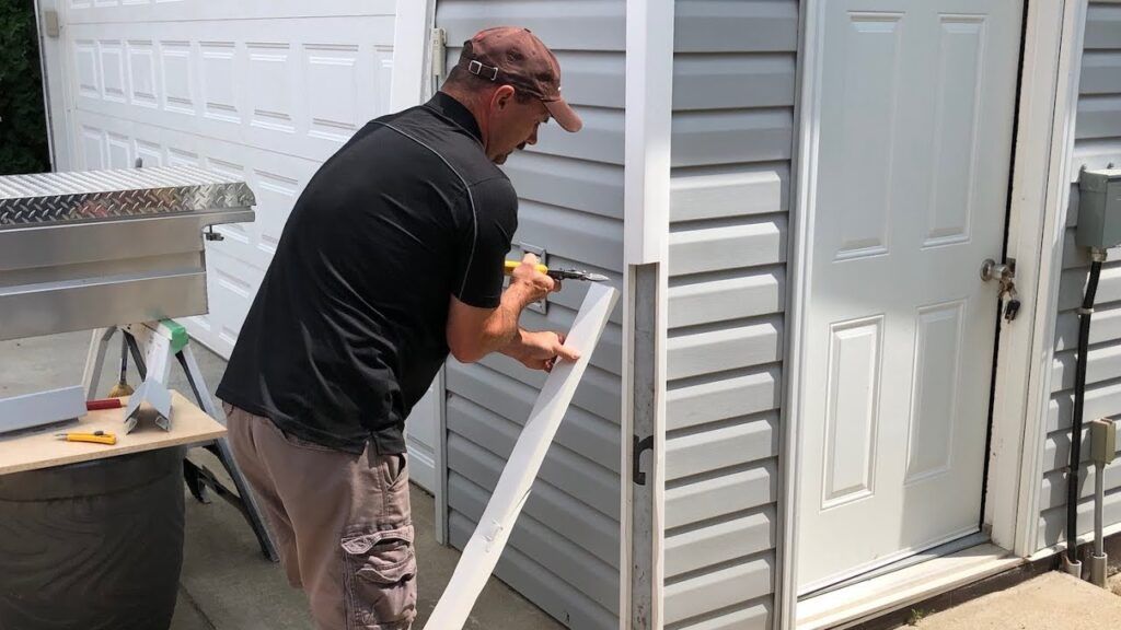 Man in cap and black shirt cutting trim on gray vinyl siding near a white door.