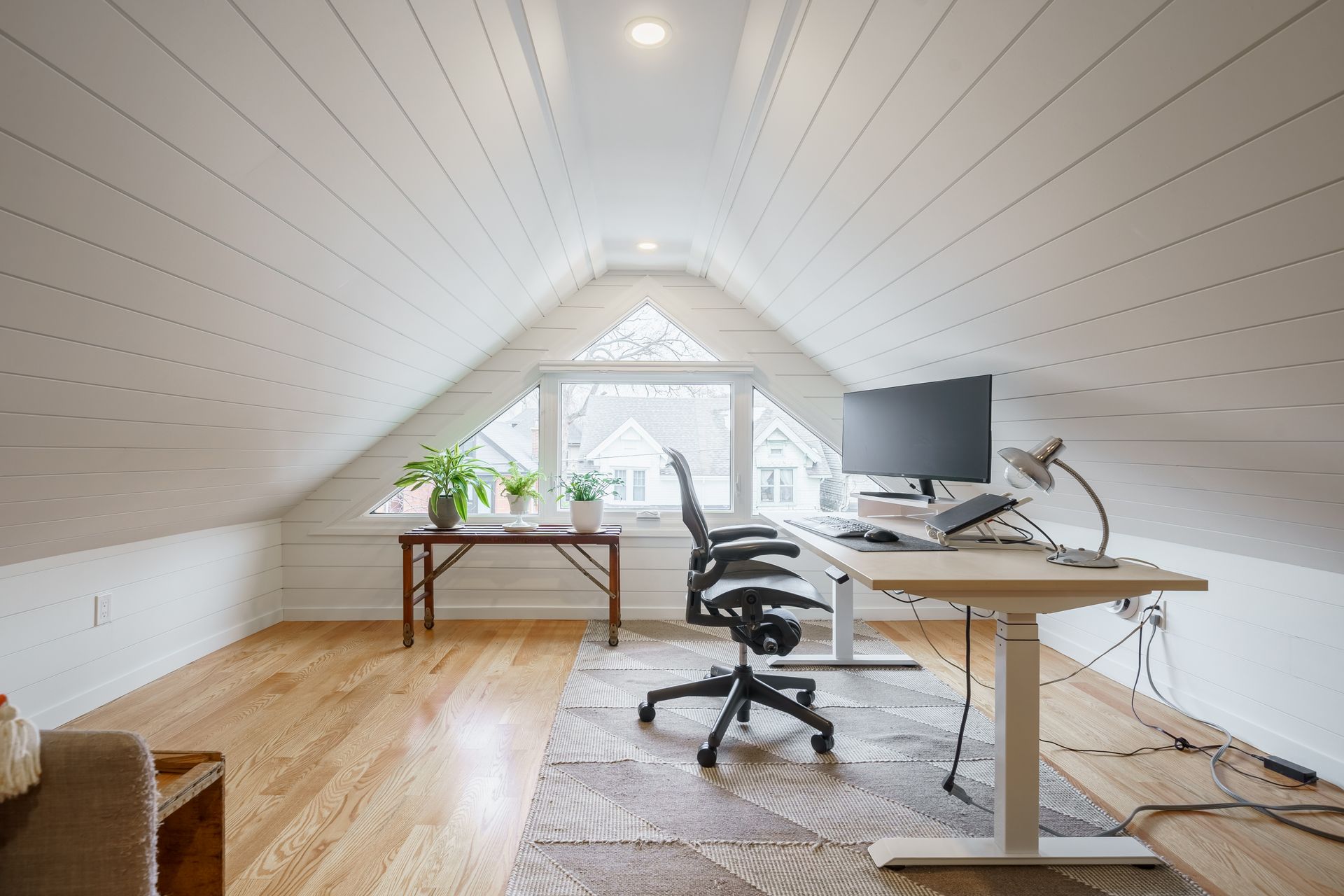 Bright attic home office with desk, monitor, chair, rug, plants, and skylight windows