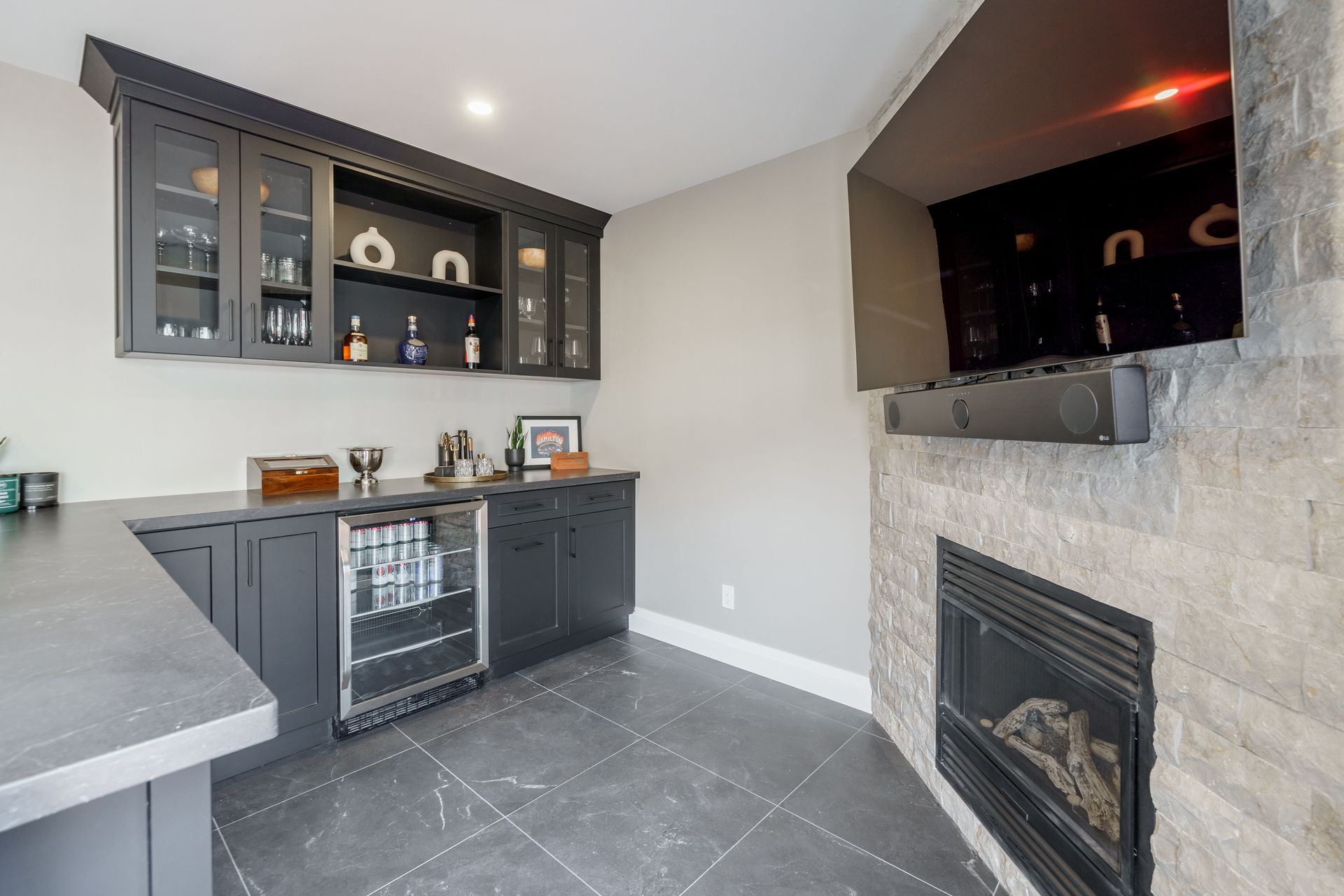 Modern kitchen with dark cabinets, gray tiled floor, and a stone fireplace on the right.
