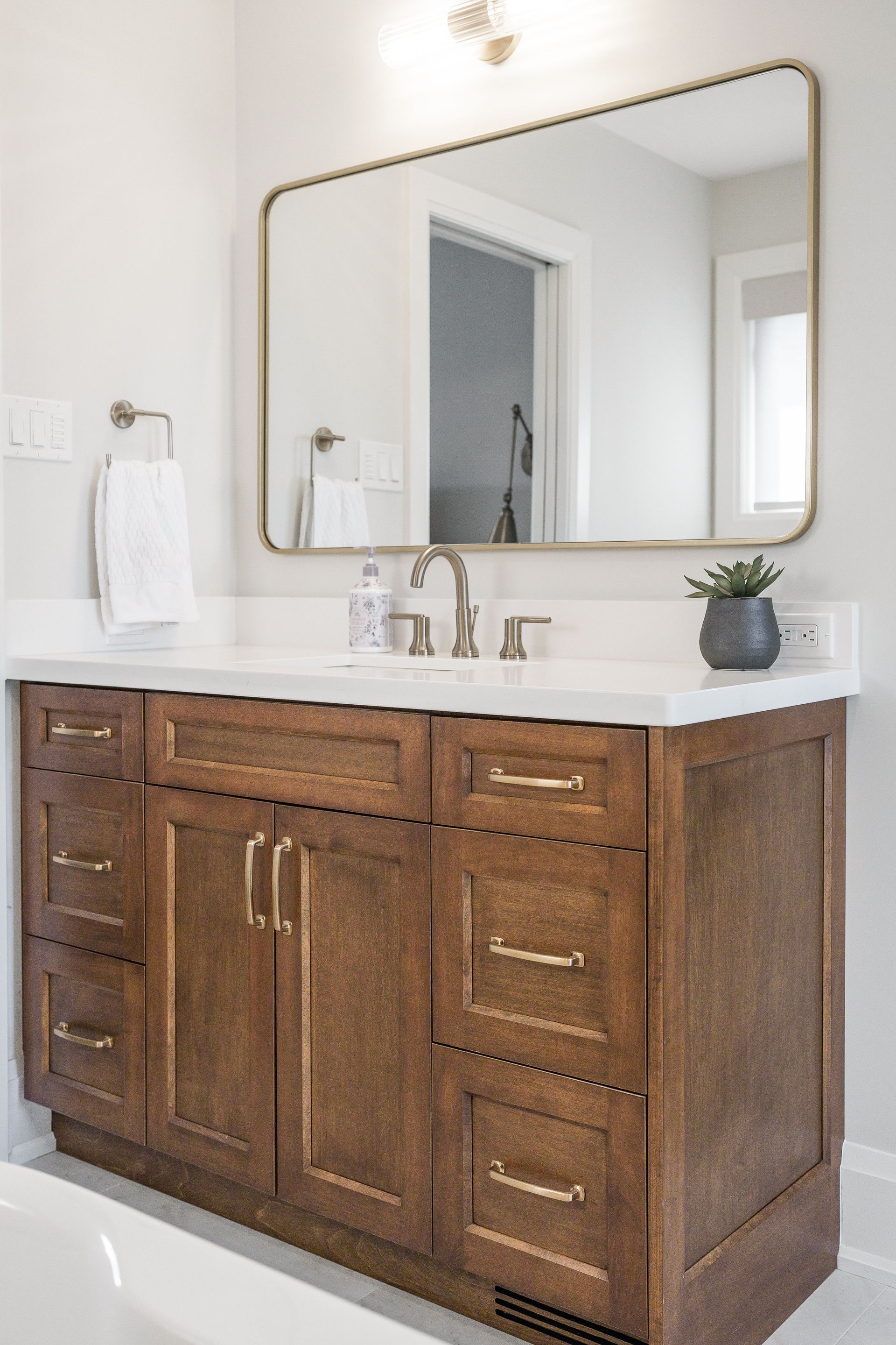 Wooden bathroom vanity with white countertop, brass faucet, mirror, and small potted plant.