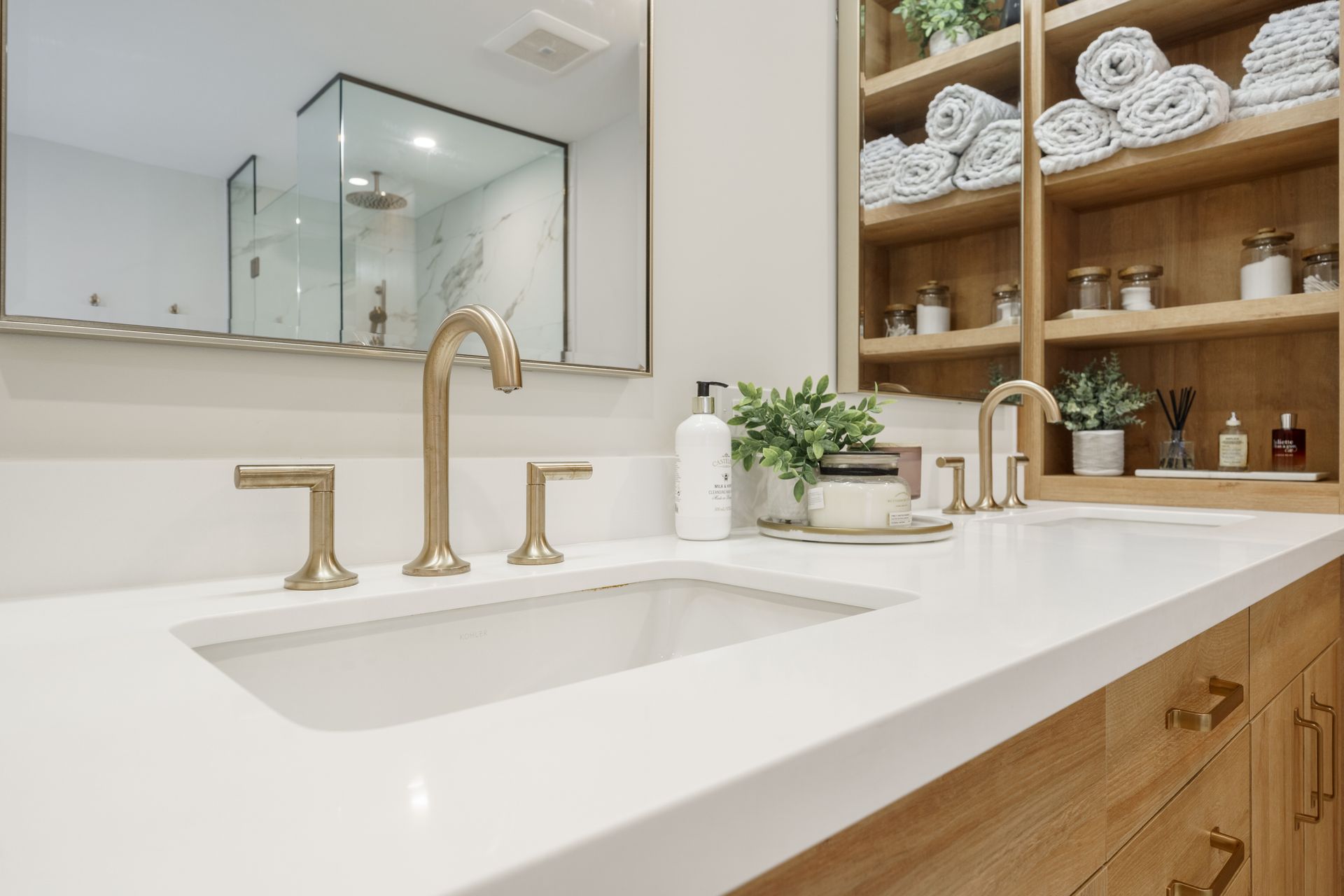 Modern bathroom vanity with white countertop, gold faucet, mirror, and wooden shelves with towels and decor