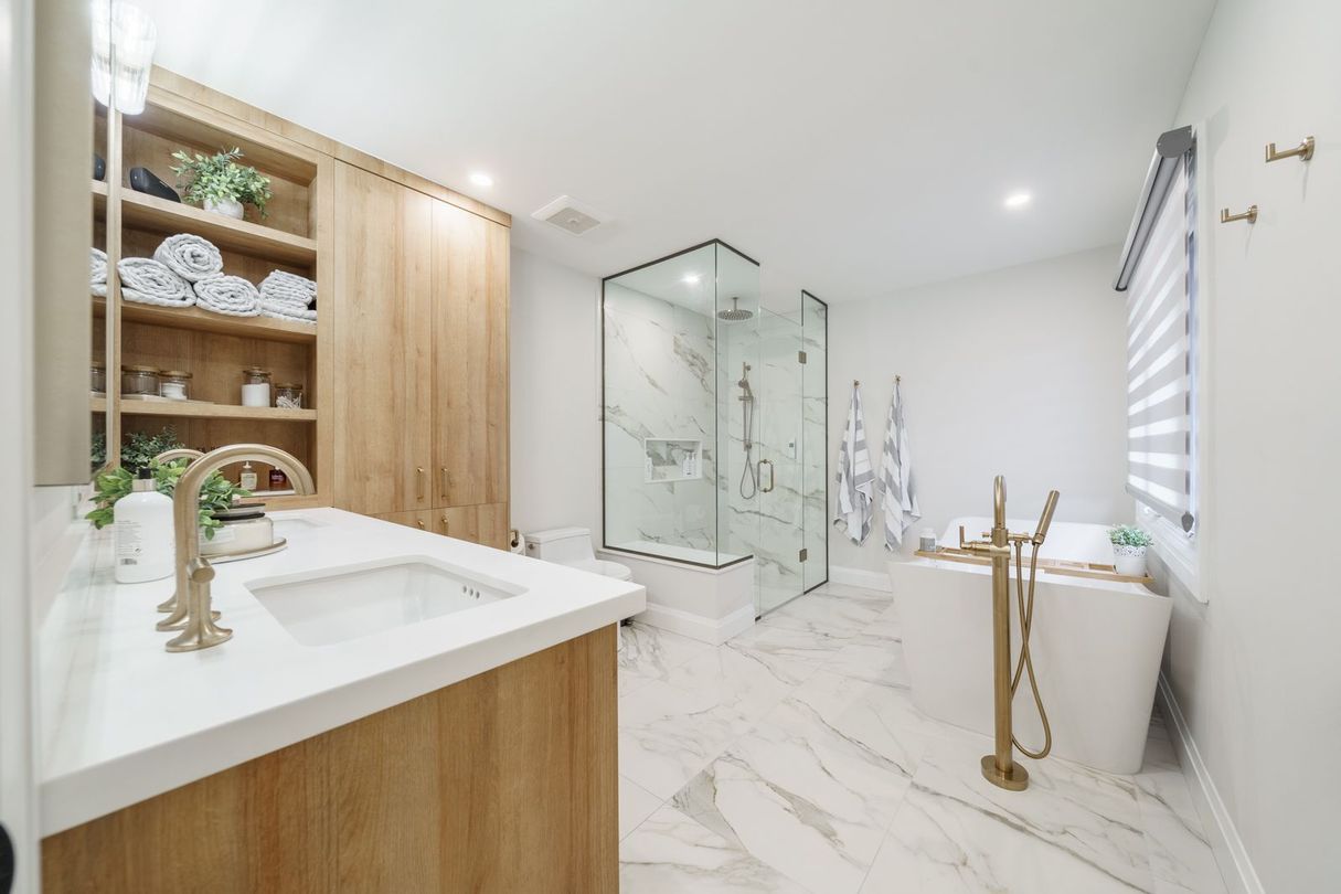 Bright modern bathroom with marble floor, wooden vanity, glass shower, and brass fixtures.