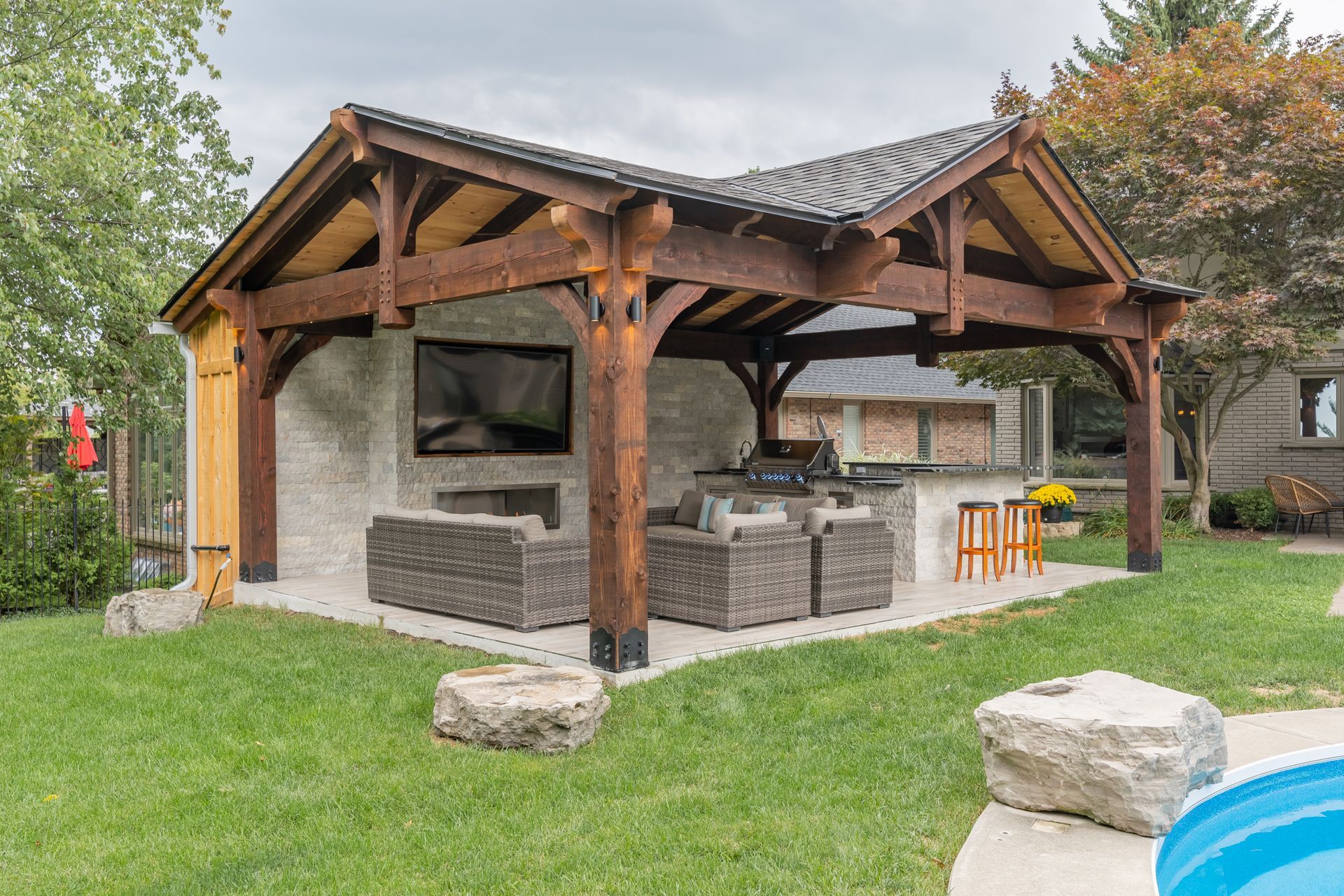 Wooden poolside pavilion with stone bar, grill, and stools on a grassy lawn