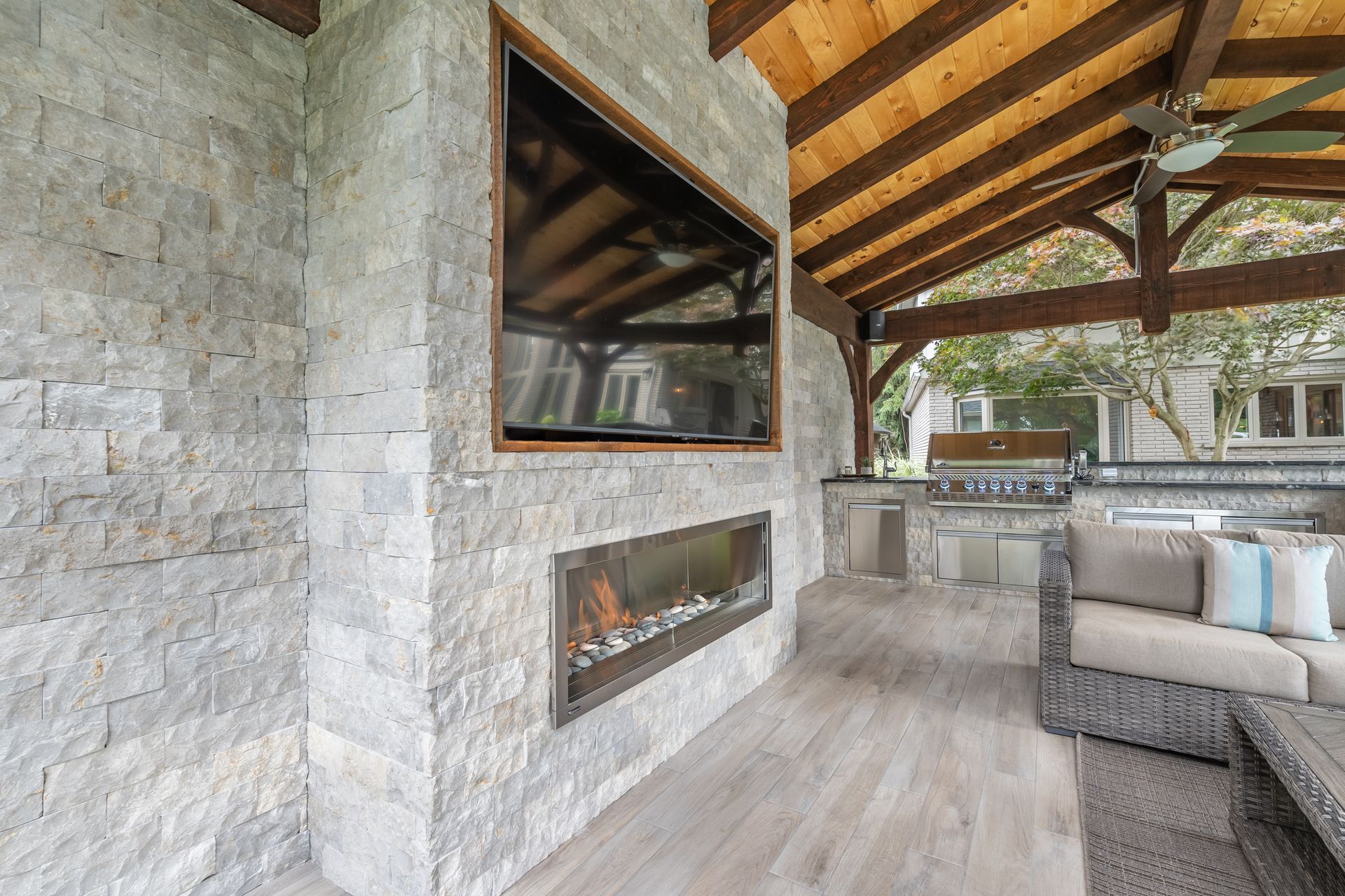 Stone outdoor patio with modern fireplace, wood-beamed ceiling, and gray sectional seating