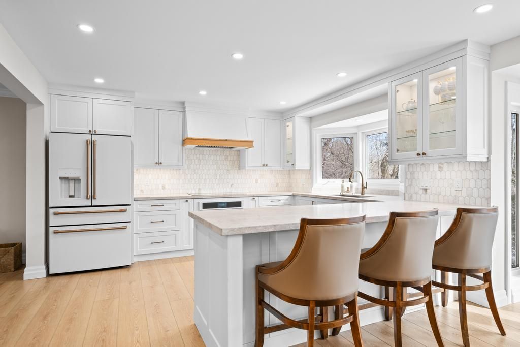 Bright modern white kitchen with island, stainless fridge, and four bar stools by a window