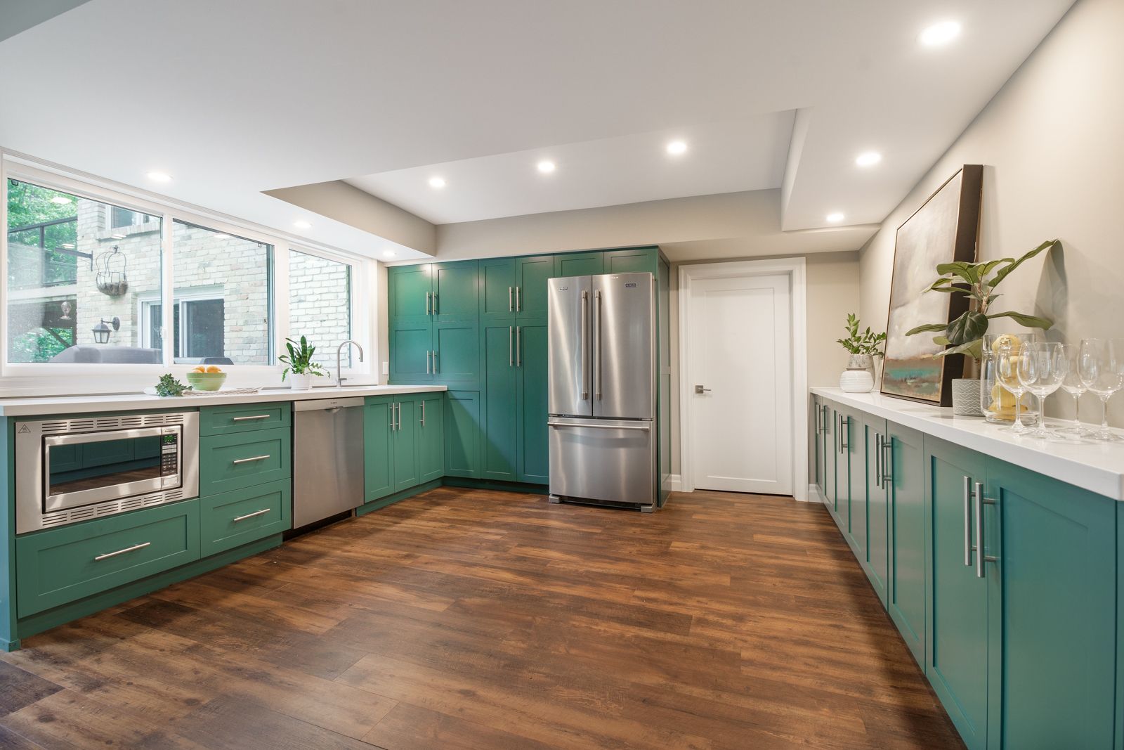 Bright teal kitchen with stainless steel appliances, white counters, wood floor, and a large window.