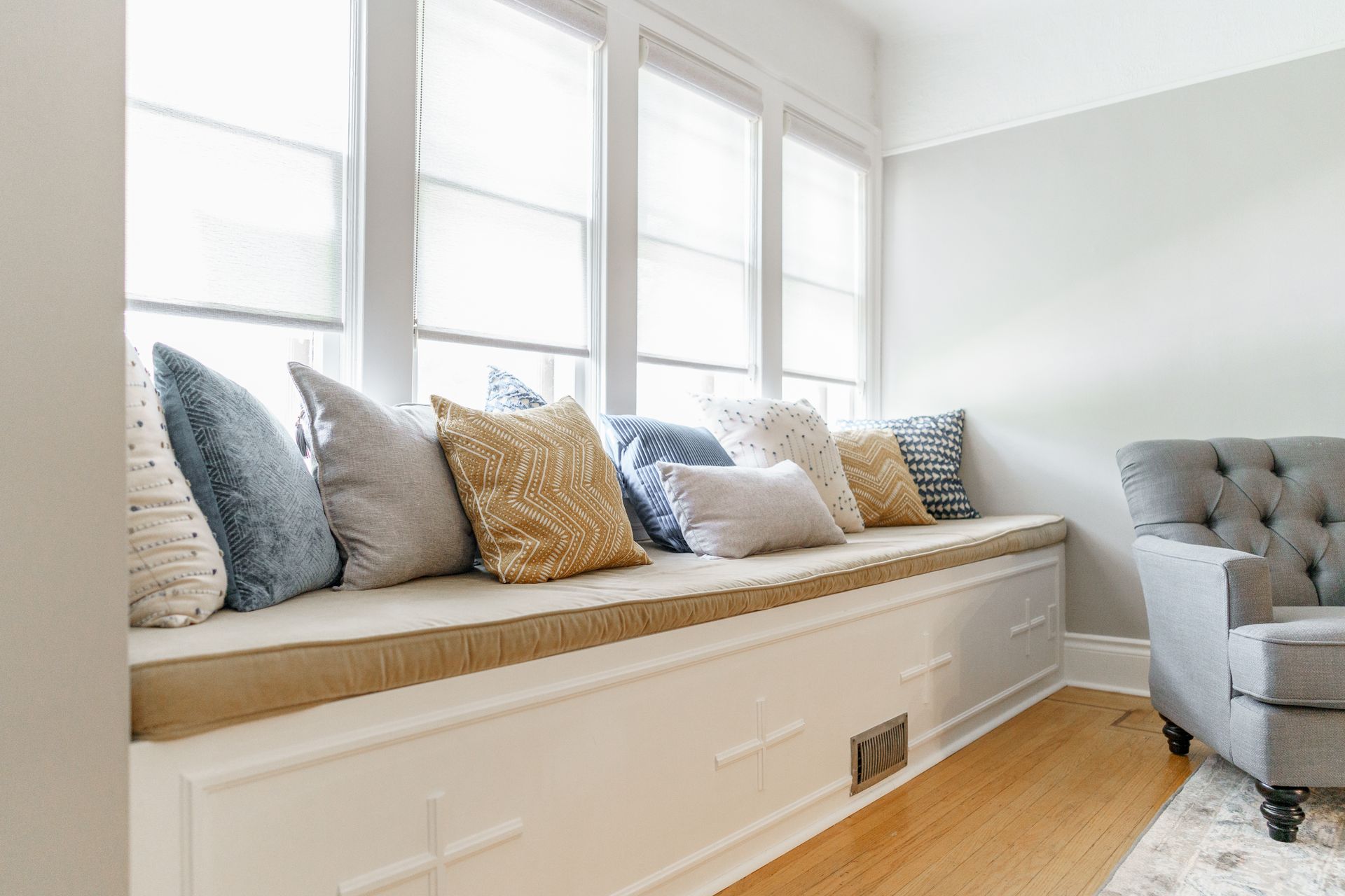 Bright window seat with white storage bench, patterned pillows, and a gray tufted chair in a sunlit room