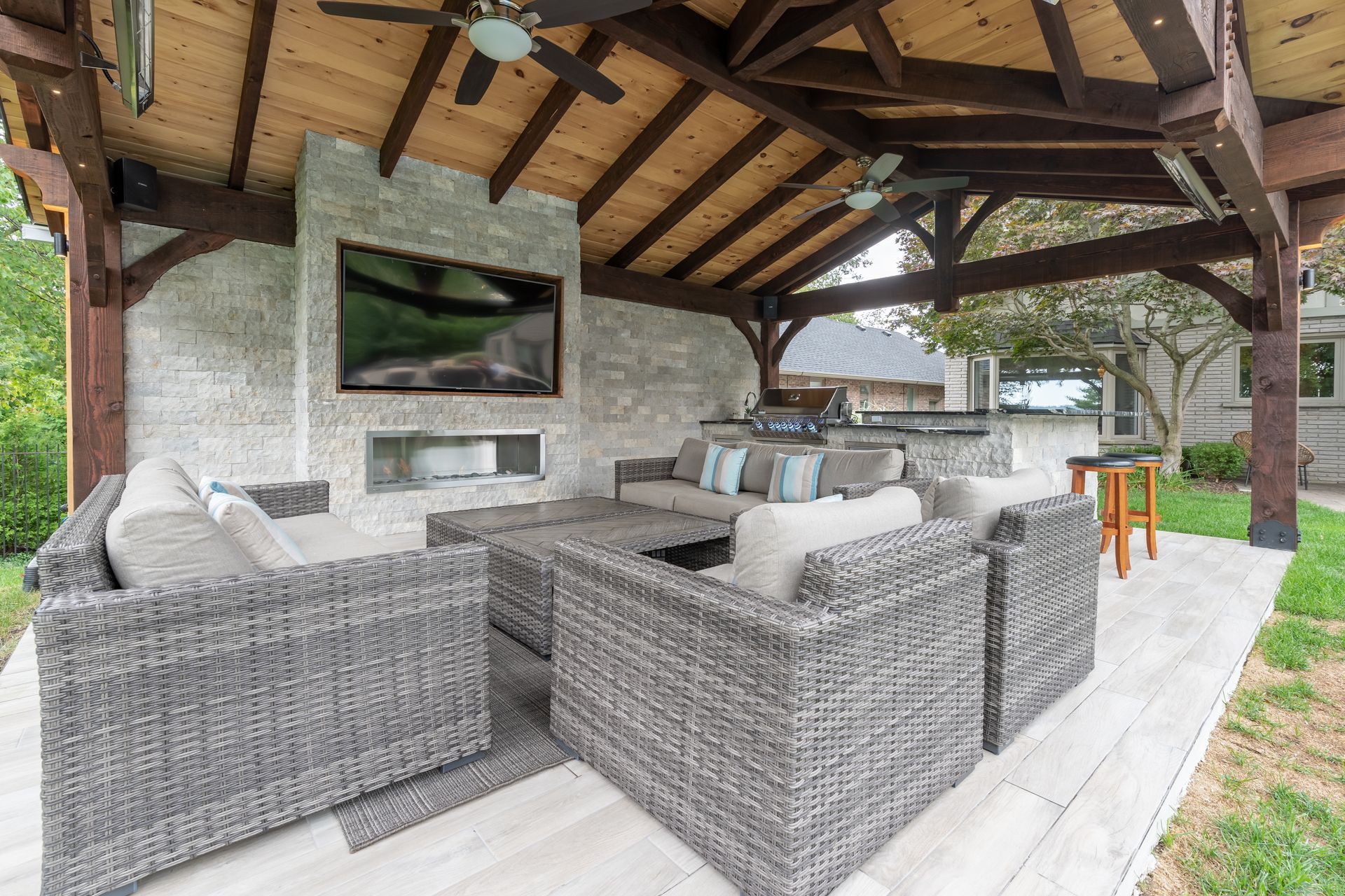Covered outdoor patio with wicker seating, wall-mounted TV, and bar under a wooden beam roof