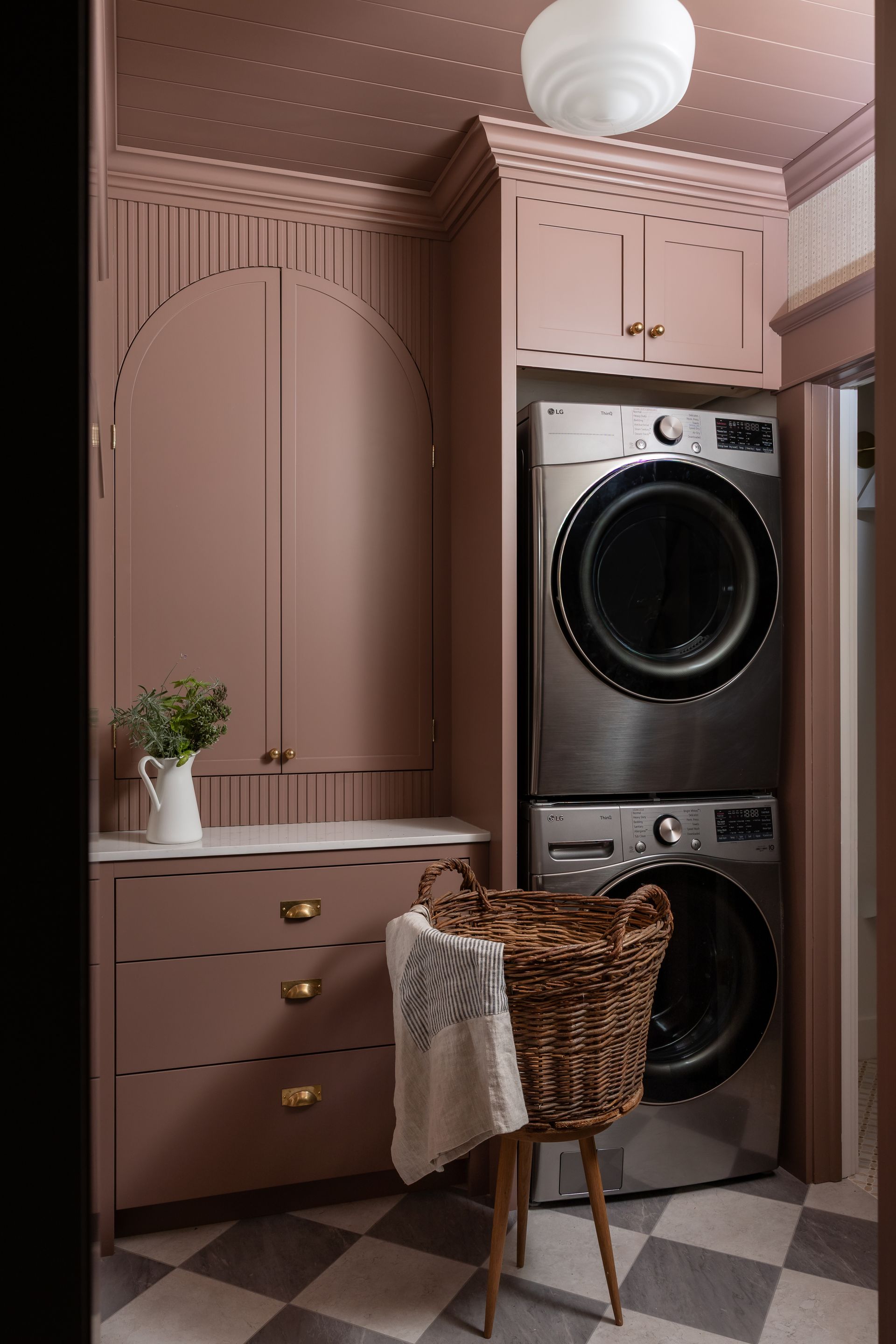 Pink laundry room with stacked washer and dryer, built-in cabinets, wicker hamper, and checkered floor