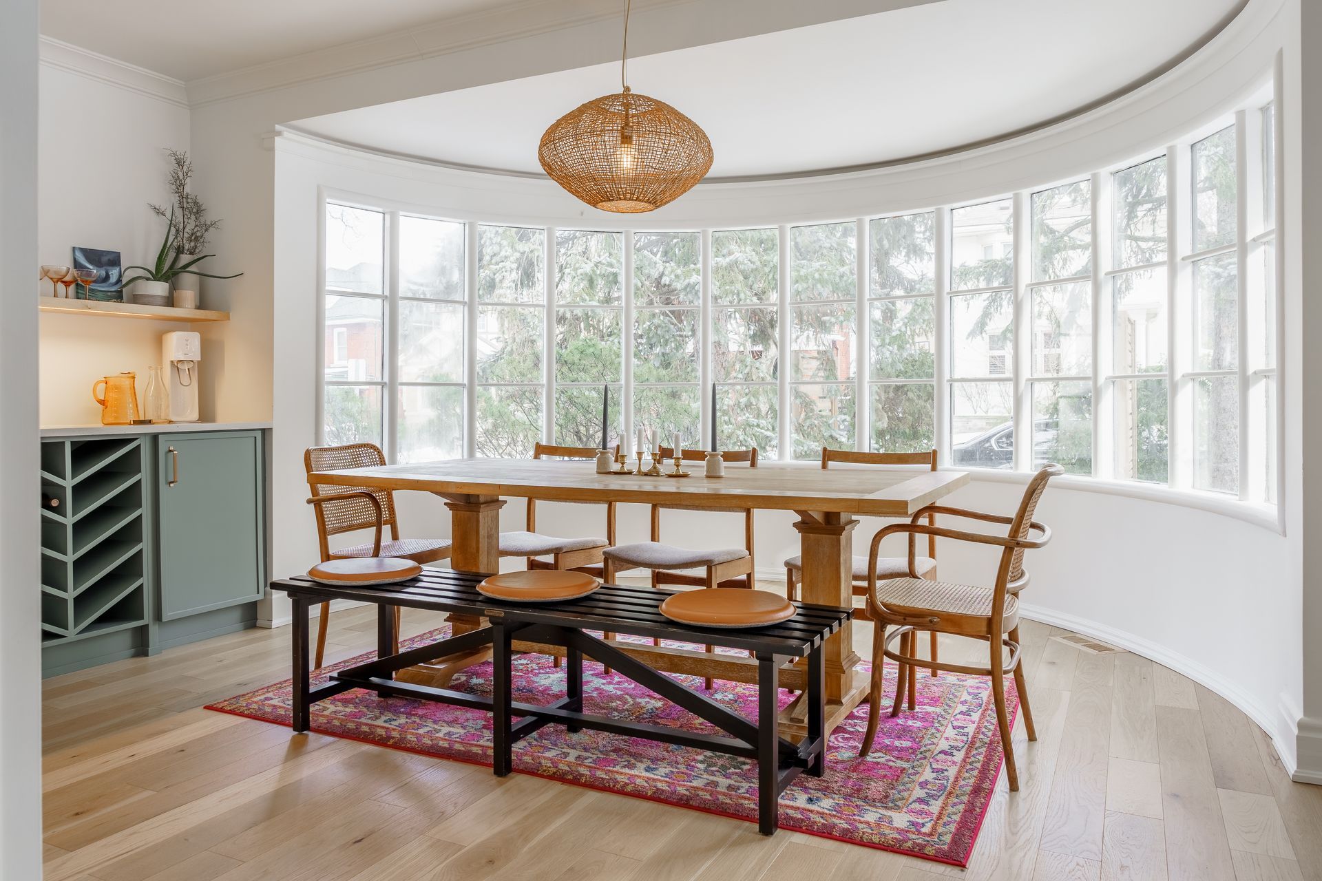 Bright sunroom with curved windows, round wooden table, wicker chairs, and patterned rugs
