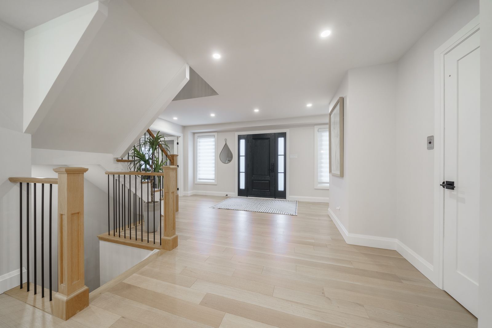 Bright empty hallway with recessed lights, light wood floors, staircase railing, and black front door at the end