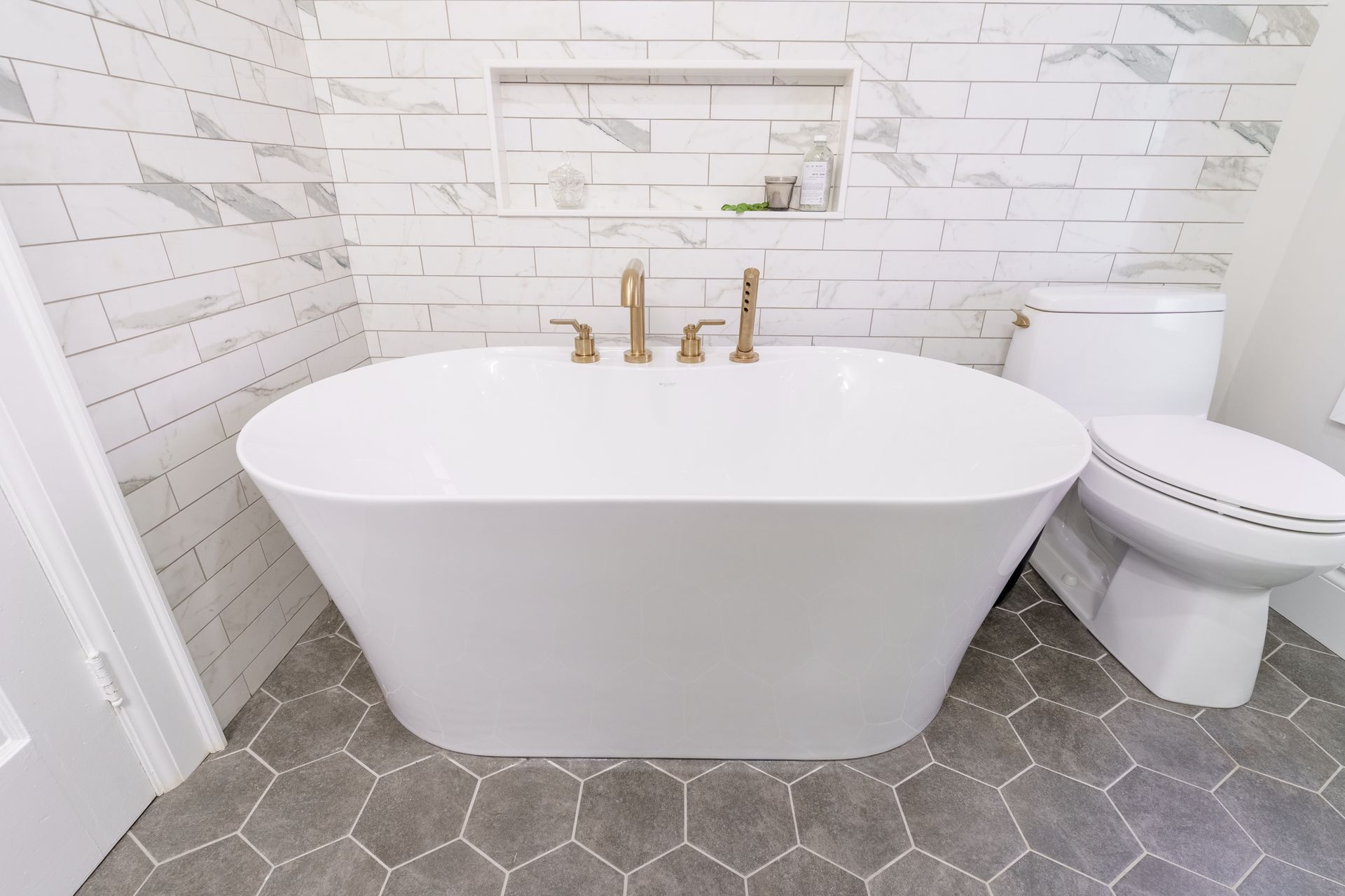 Modern bathroom with white freestanding tub, gold fixtures, hex tile floor, and toilet