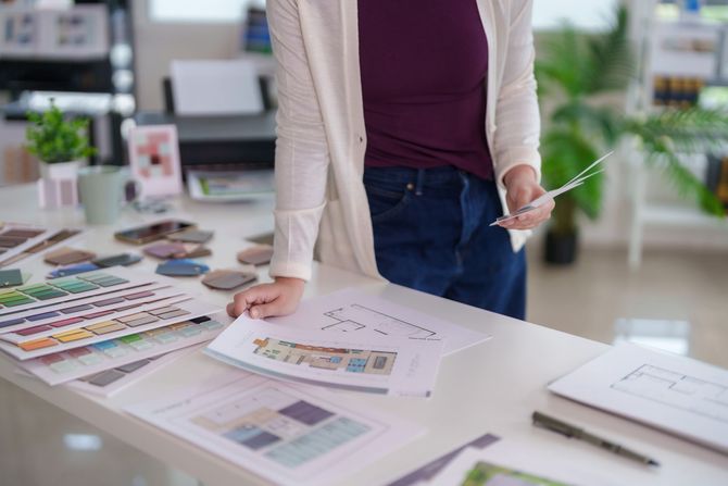 Person standing at a desk reviewing design color swatches and print layouts in a bright studio