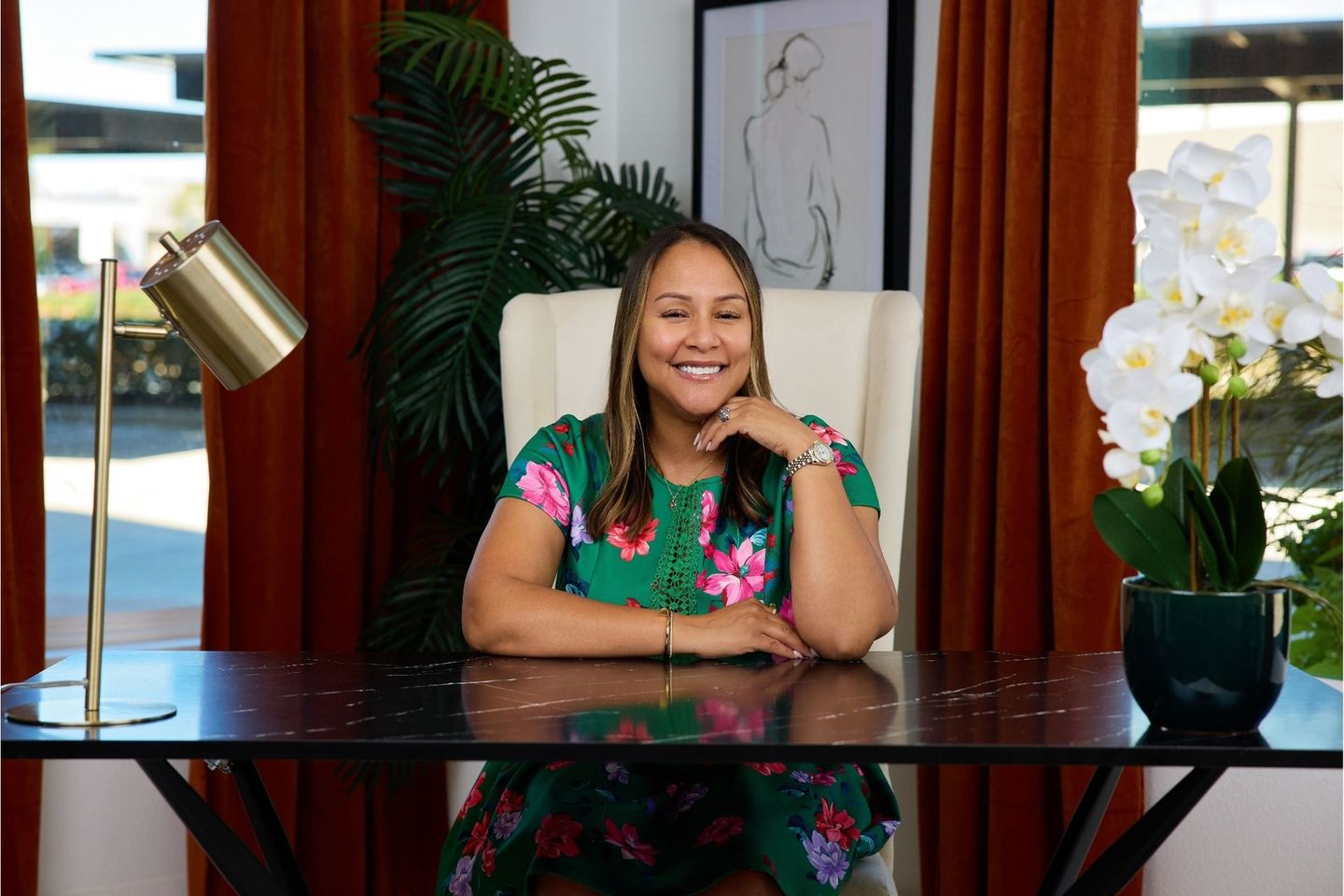 Smiling woman in a floral dress seated at a desk in a bright room with plants and curtains