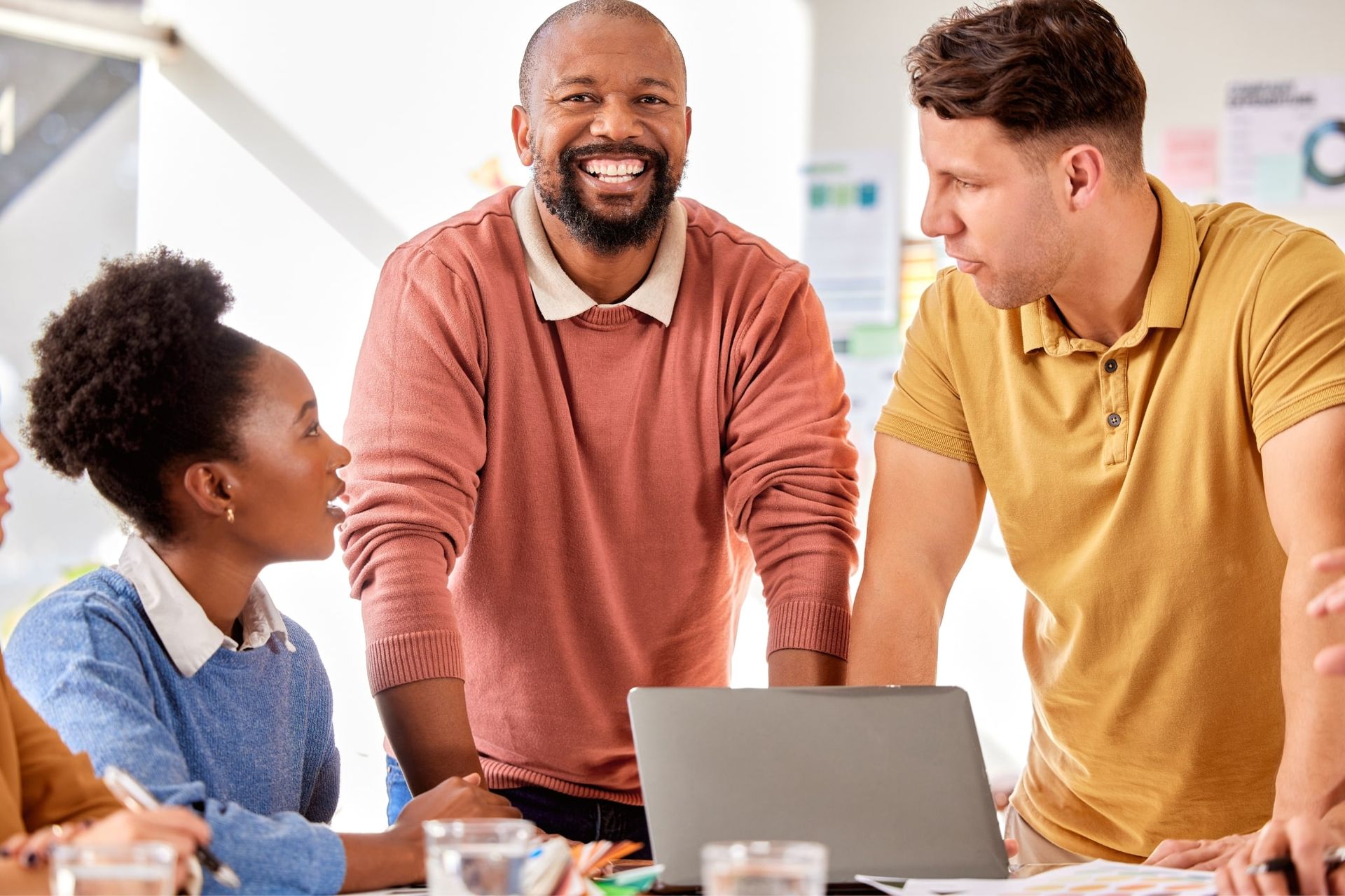 Three coworkers smiling and talking around a laptop in a bright office meeting room