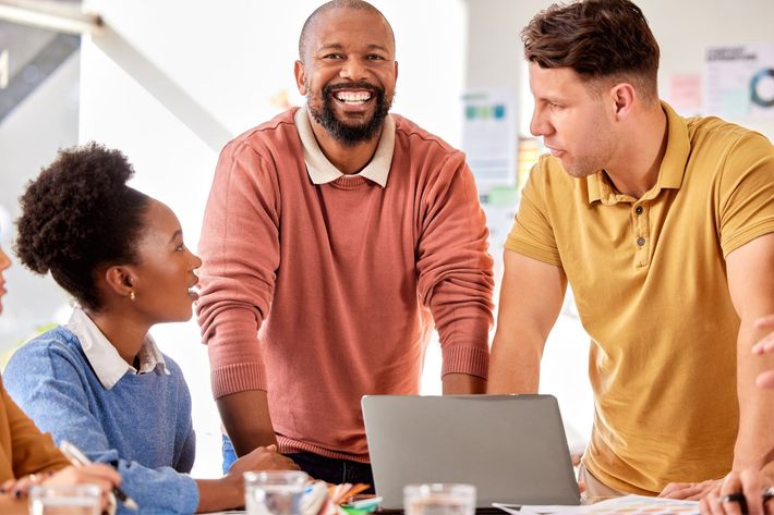 Three coworkers smiling and talking around a laptop in a bright office meeting room