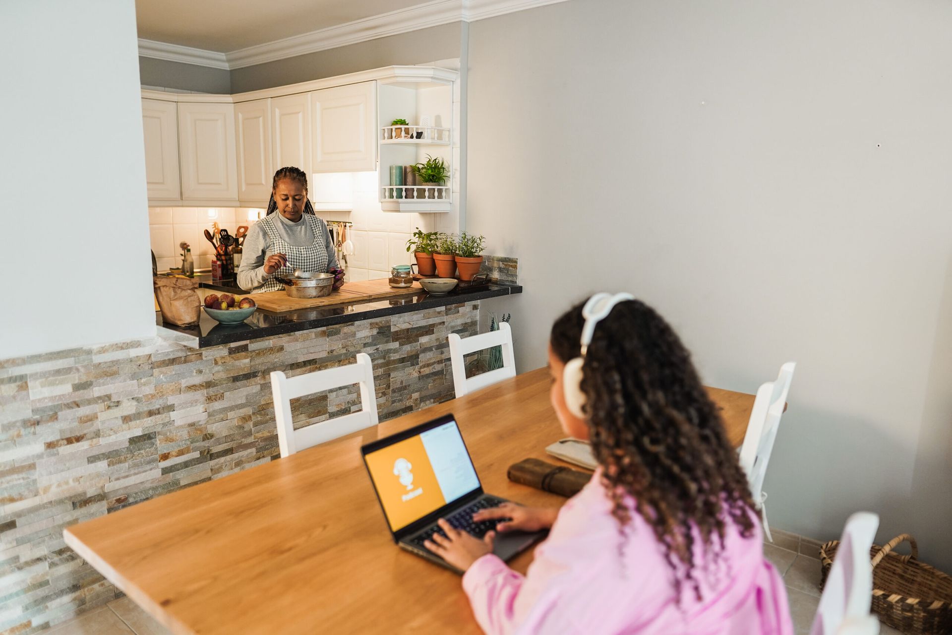 Woman using a laptop at a dining table while another person cooks in an open kitchen.