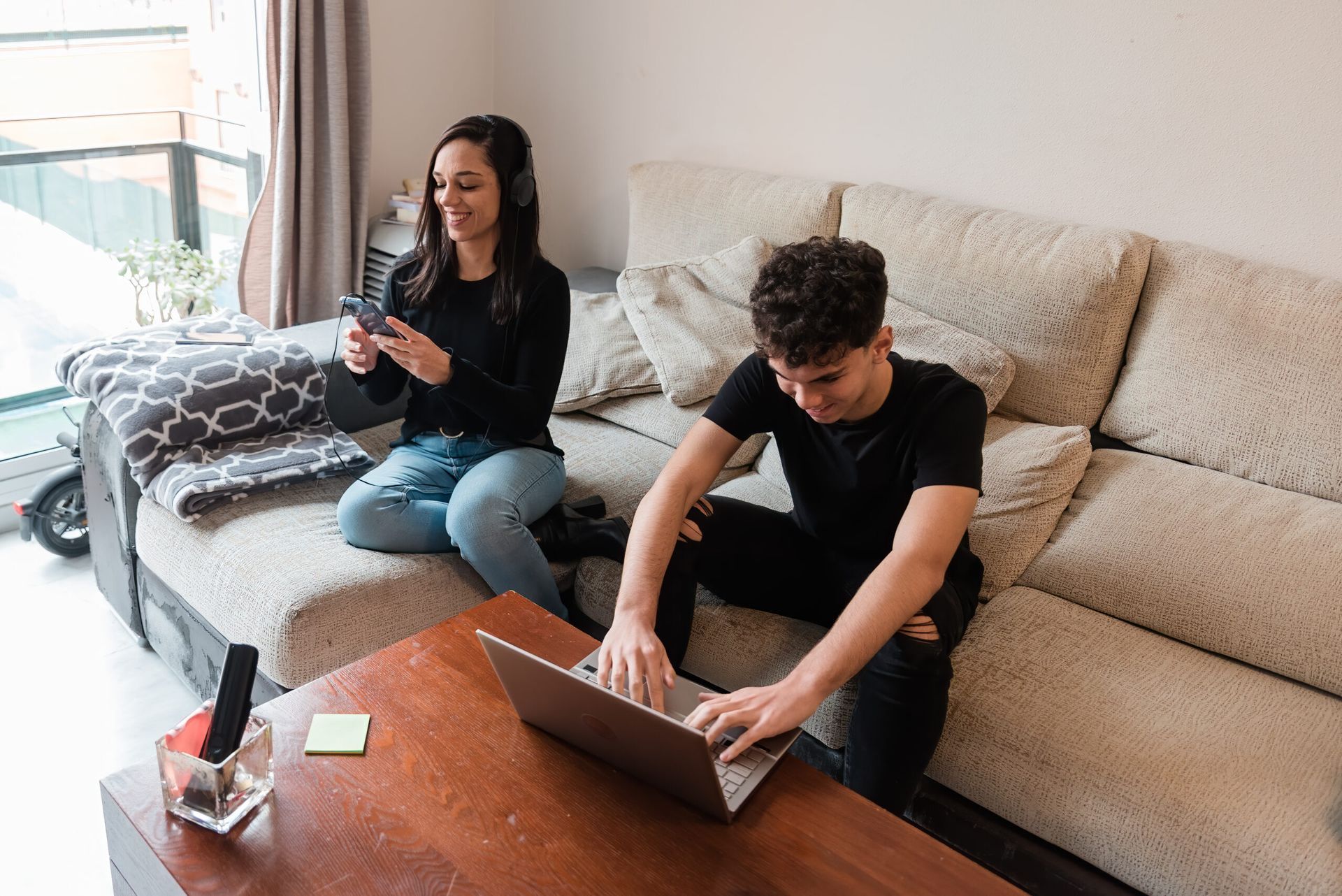 Two people on a couch, one using a laptop and the other holding a drink in a bright living room