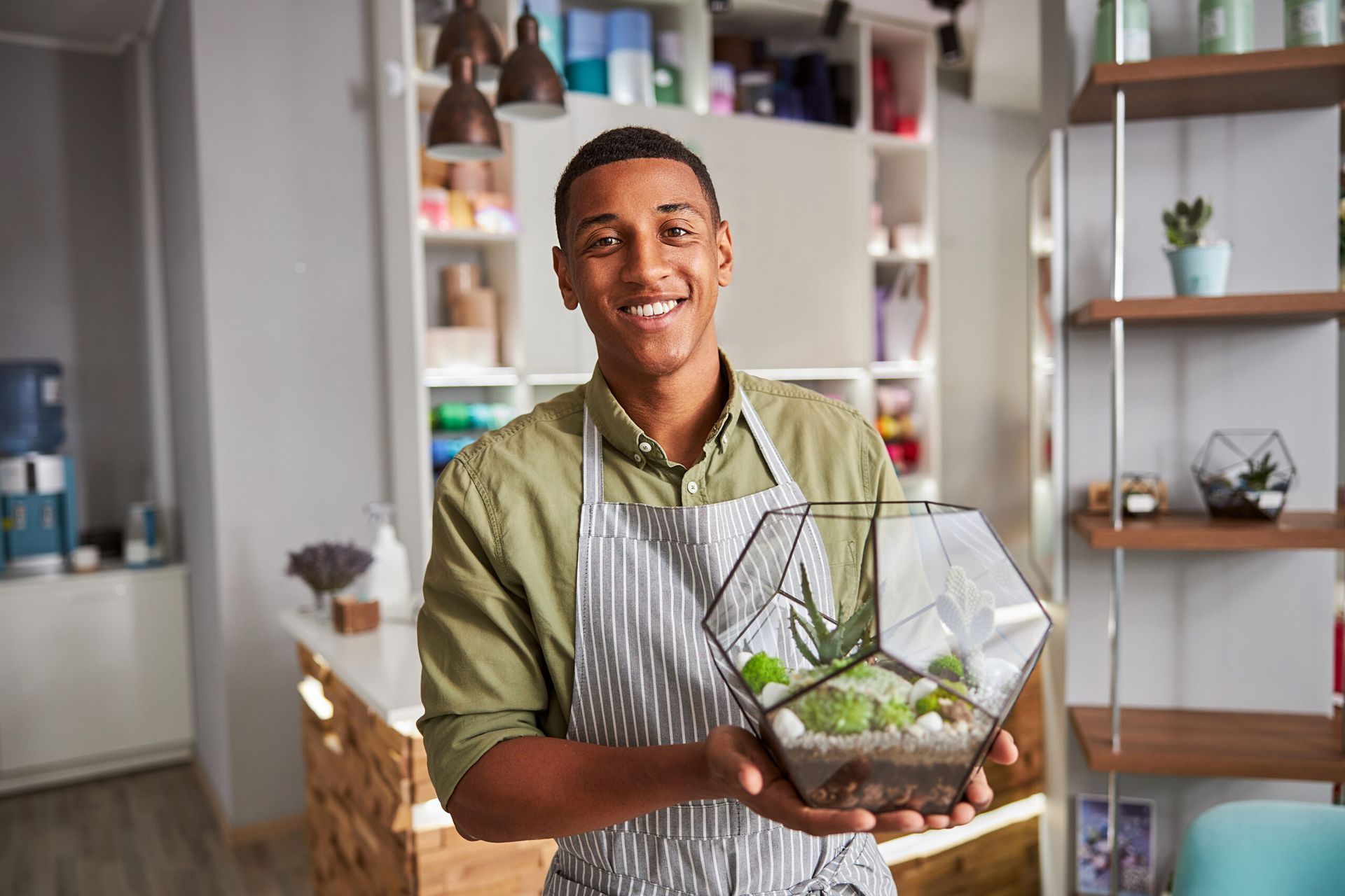 Smiling person holding a glass terrarium with plants in a bright room with shelves and decor