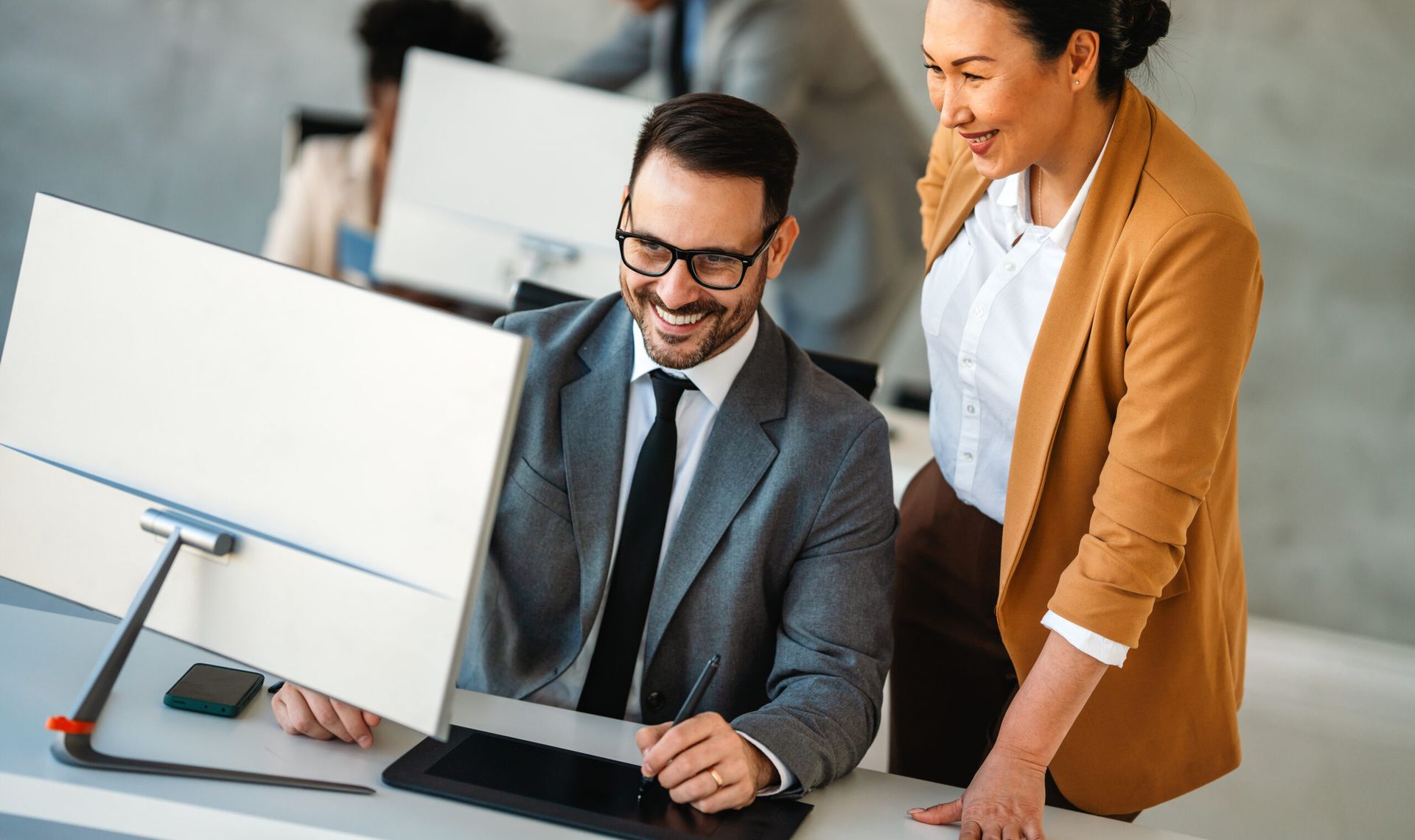 Two coworkers smiling at a desk with a computer in a bright office