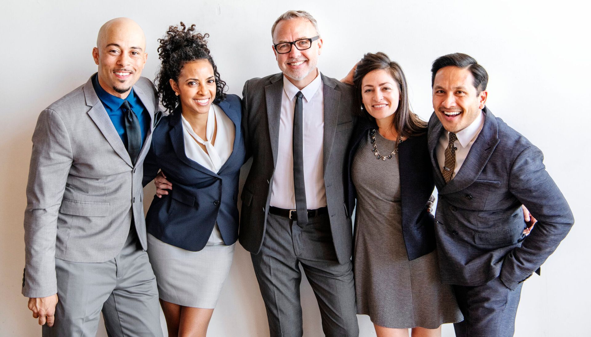 Five coworkers in business attire stand together, smiling against a plain white background.