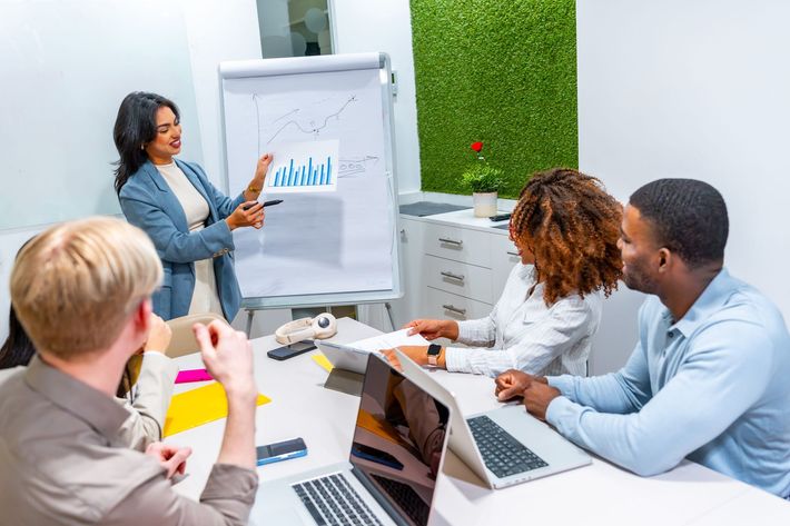 Team meeting in a bright office, presenter pointing at a chart while colleagues review documents and laptops.