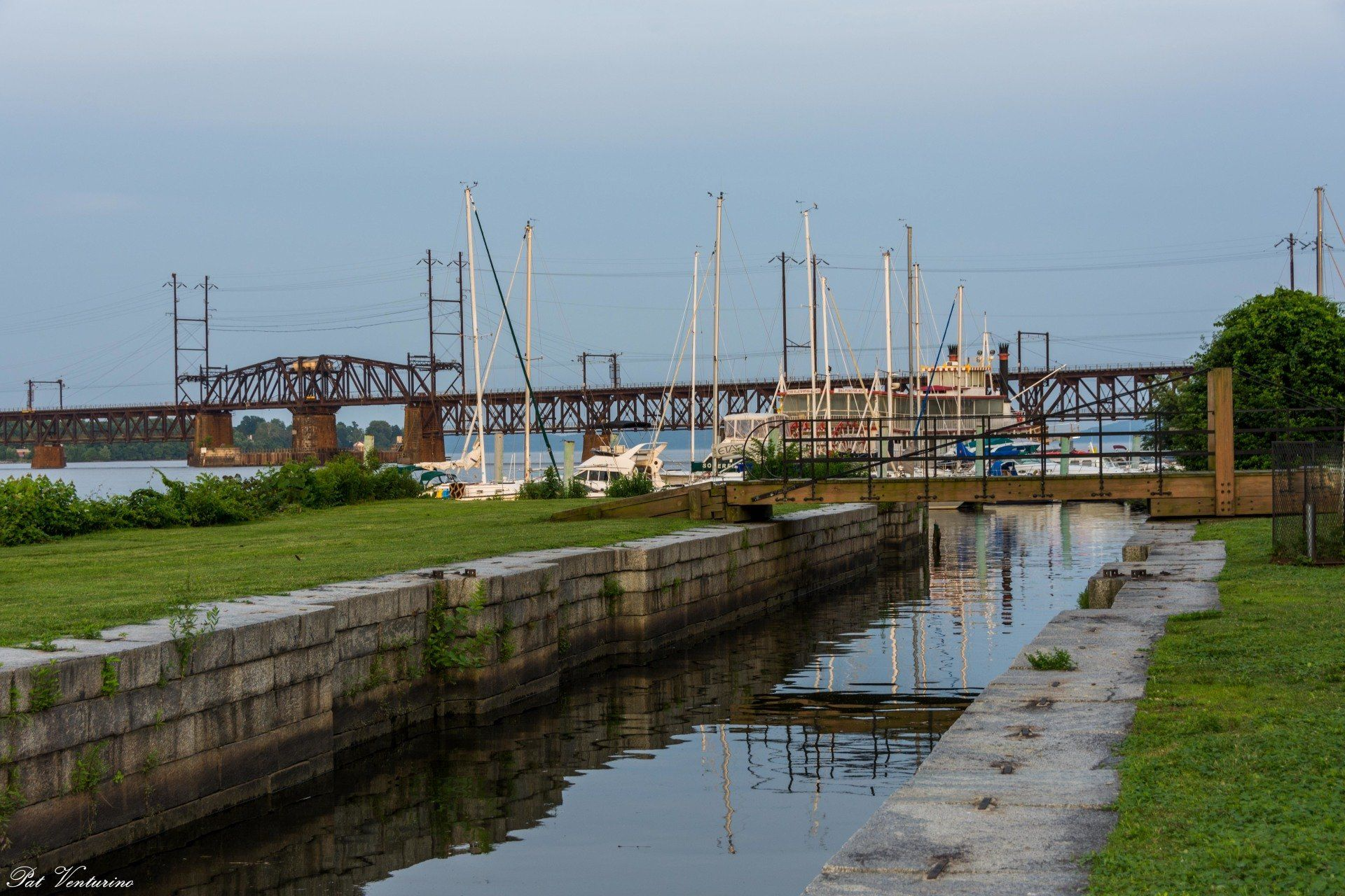 A canal with a bridge and docked sailboats in the background, at dusk.
