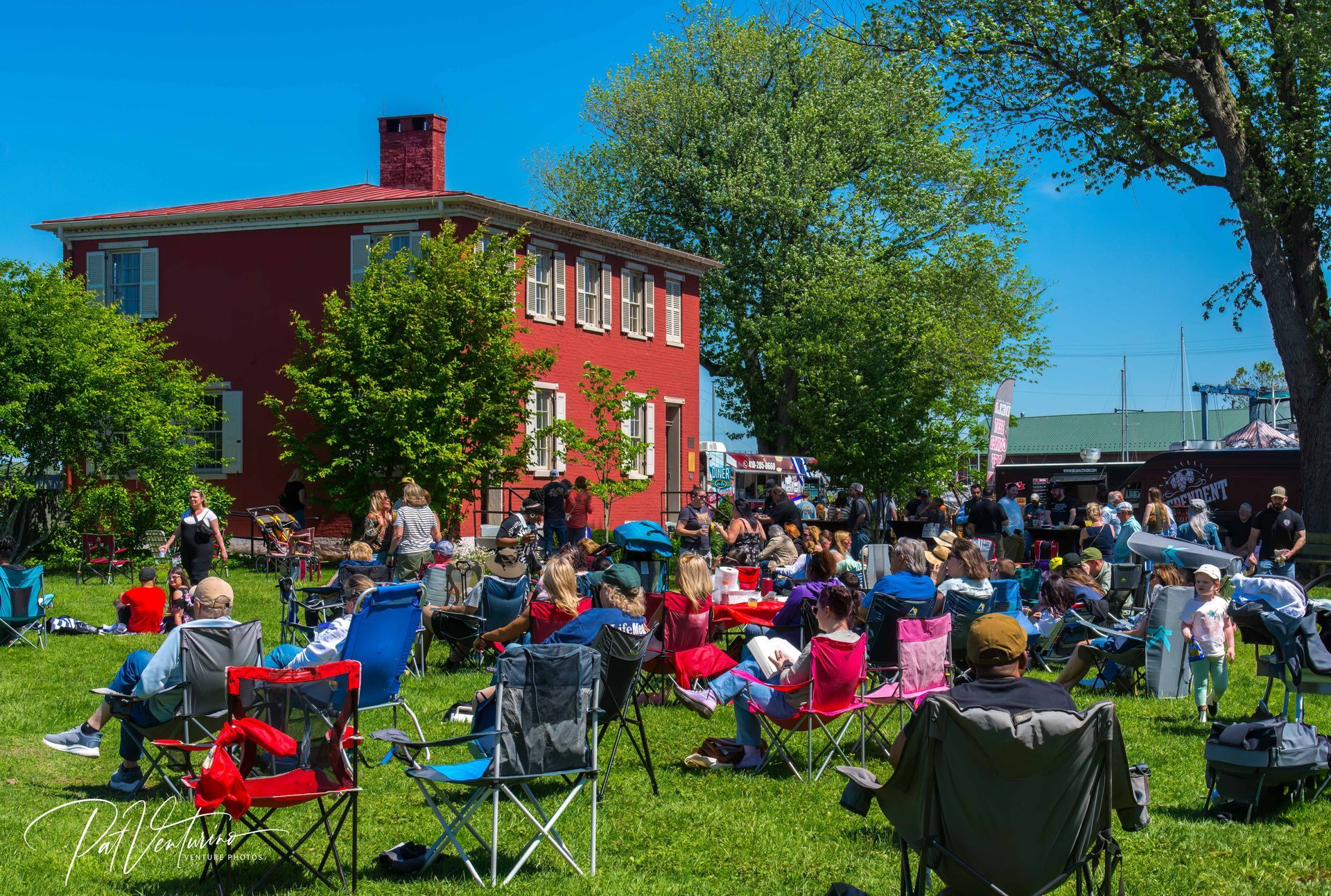 People sitting in lawn chairs at an outdoor event in front of a red building on a sunny day.