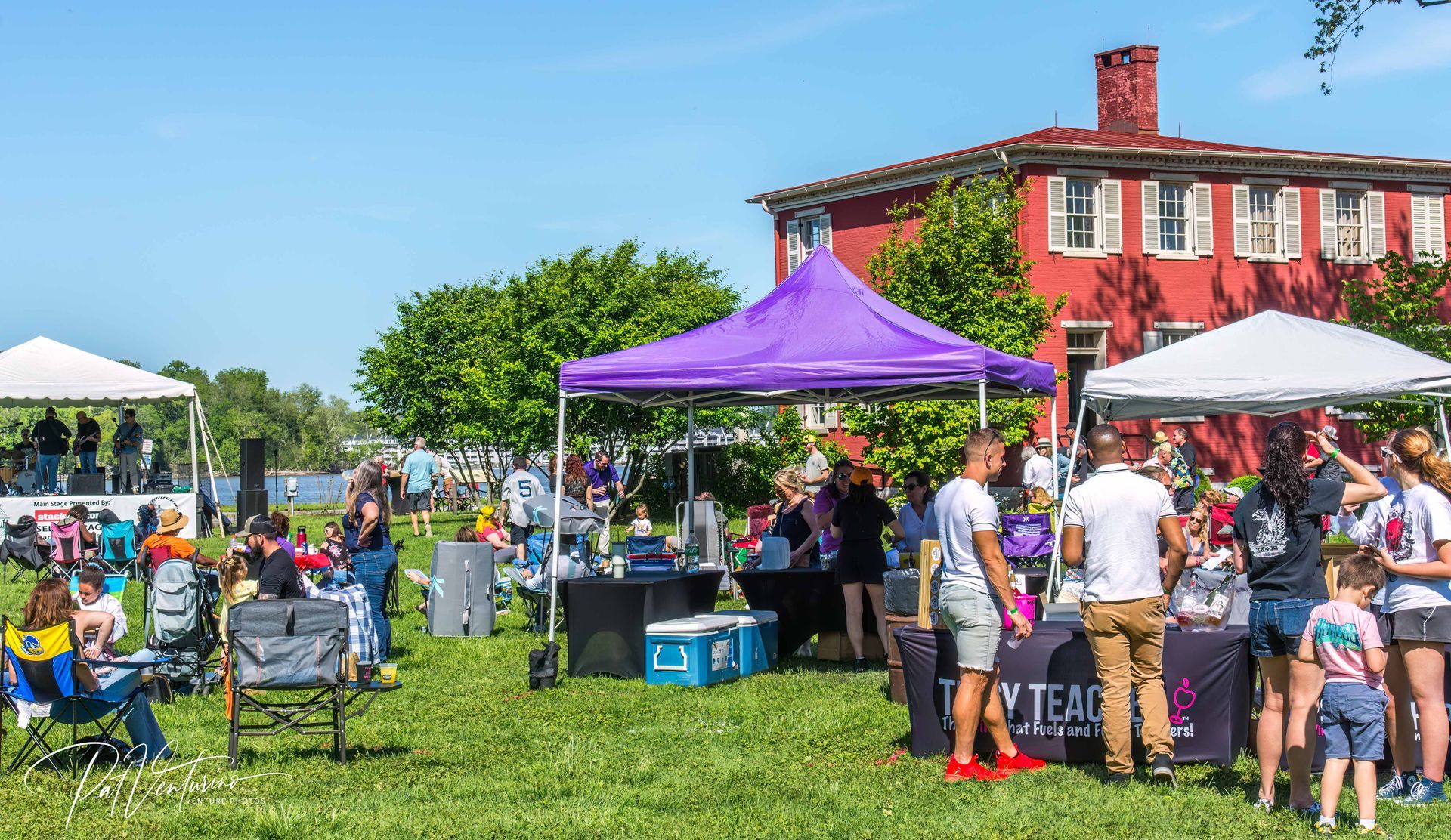 Outdoor event with vendors, people, tents, and a red building on a sunny day.
