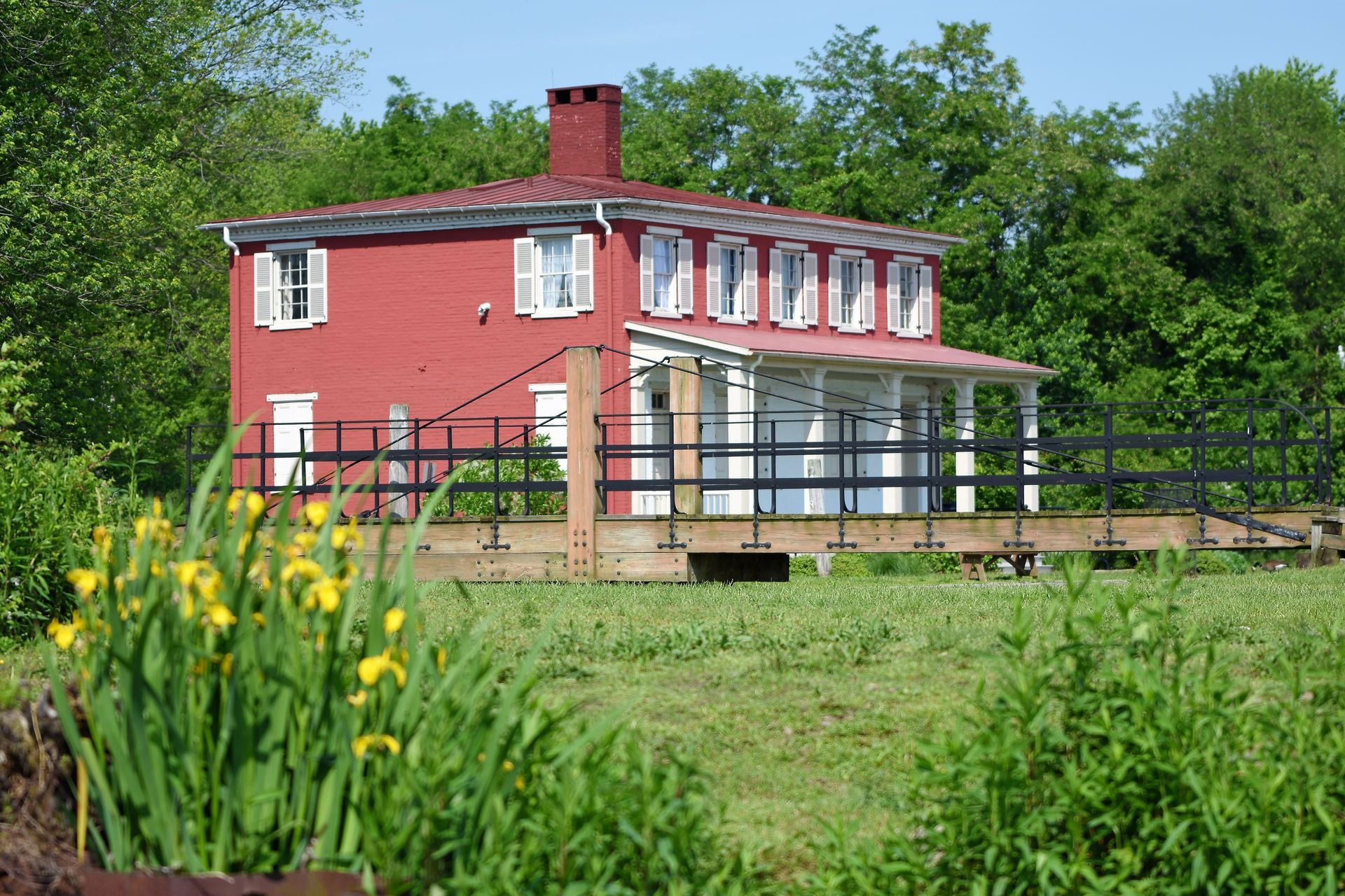 Red two-story house with white trim and shutters, surrounded by green foliage and a wooden bridge.