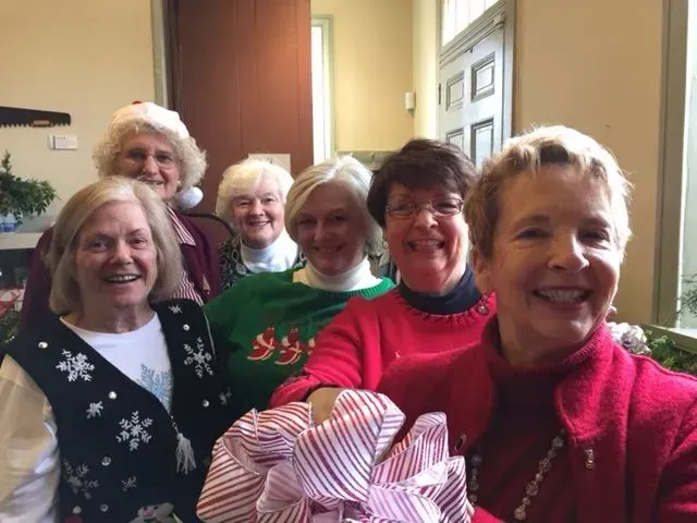 Group of smiling women in Christmas attire, holding a large wrapped gift.