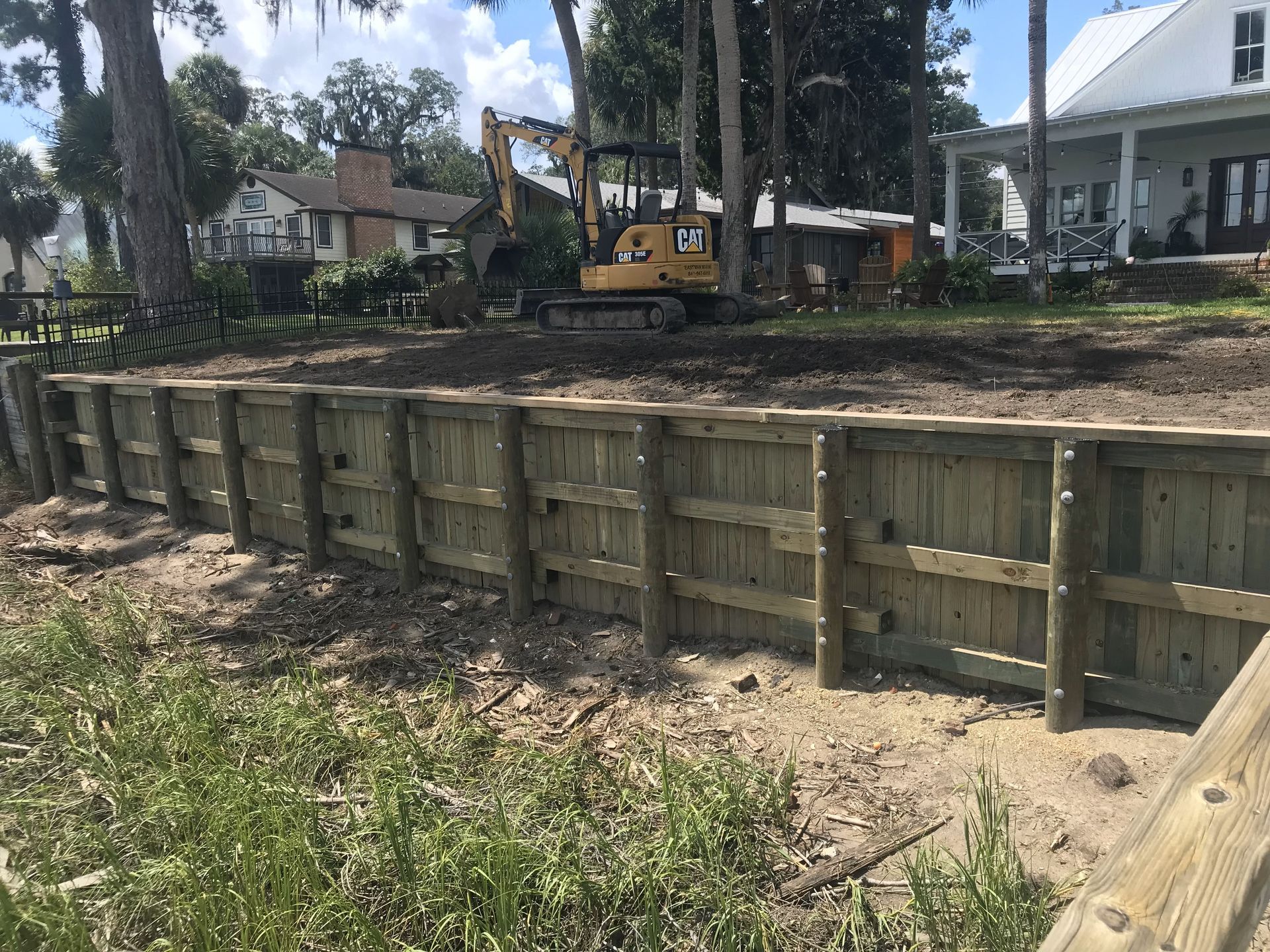 A cat excavator is working on a wooden wall in front of a house.
