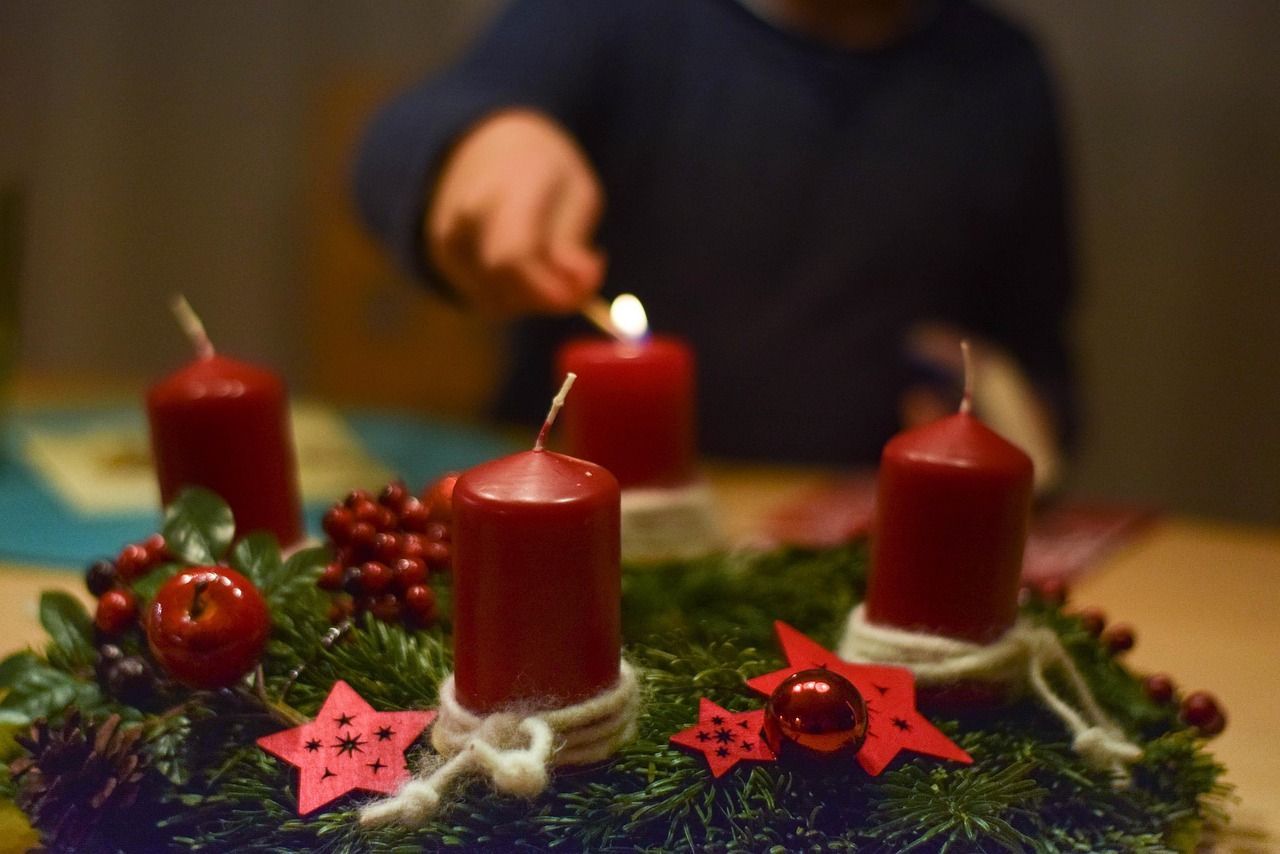 An advent wreath with four red candles. One candle is being lit by a blurry figure in the background.