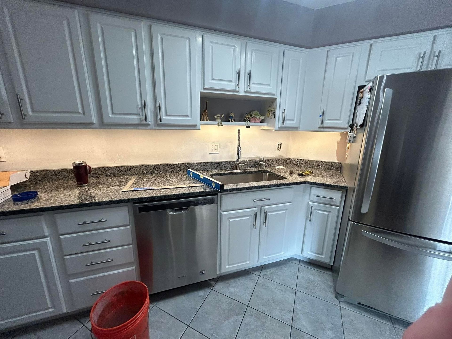 Modern kitchen with white cabinets, granite counters, stainless appliances, and a red bucket on the floor.