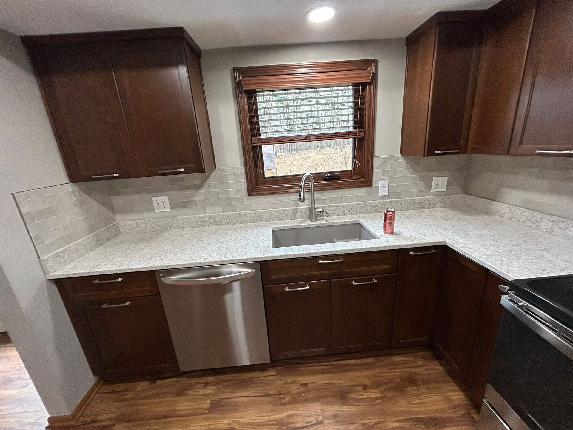 Small kitchen with dark wood cabinets, white countertops, sink under a window, and stainless steel dishwasher.