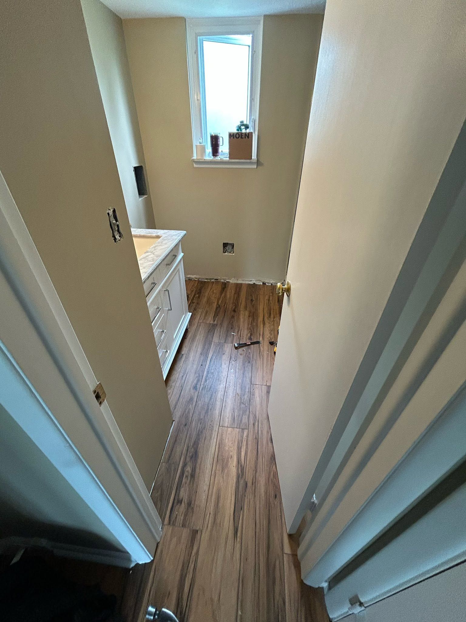 Narrow hallway into a beige bathroom with wood-look floor, white vanity, and window at the far end